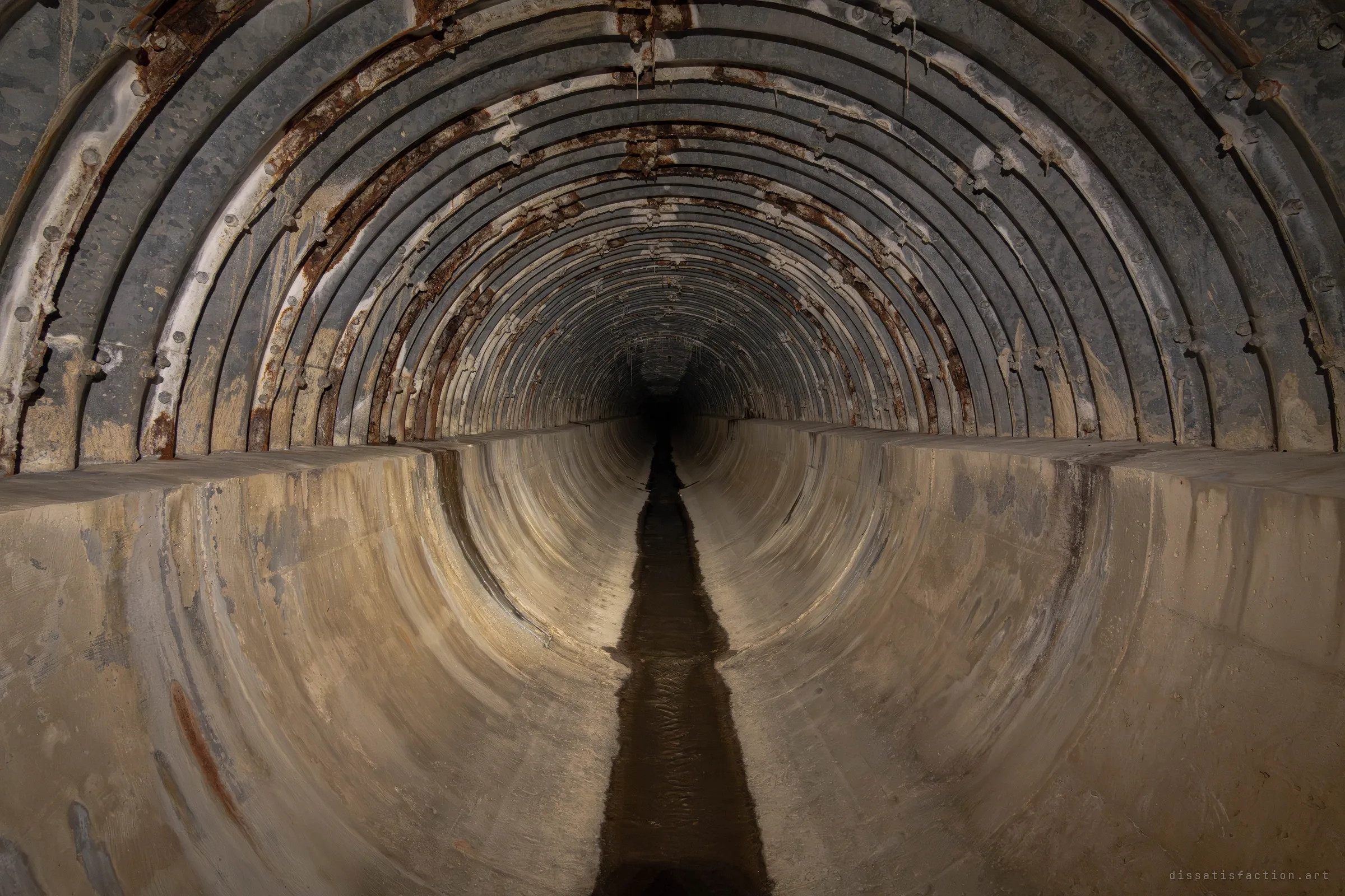 A long corrugated steel culvert, its bolted ribs streaked with rust and mineral drips, narrows into darkness above a smooth concrete channel. A thin line of dark water runs down the centre of the floor.
