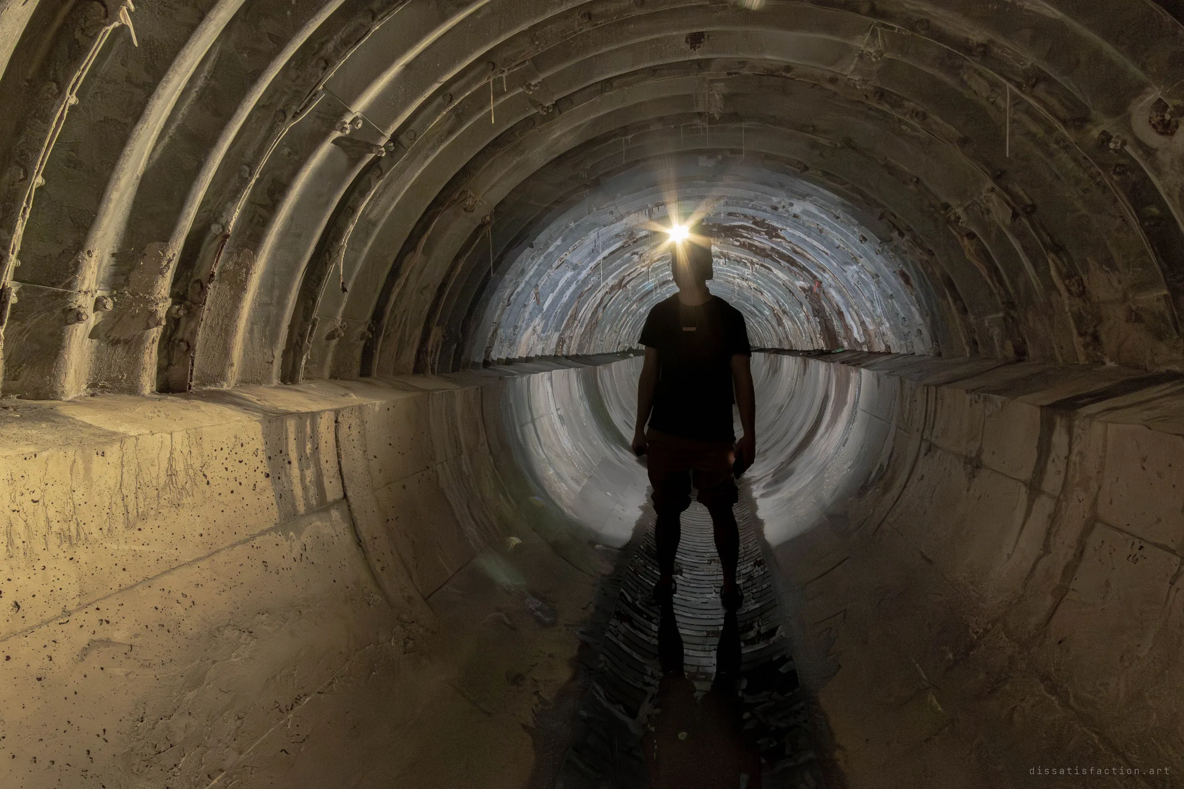 A silhouetted explorer with a glowing headlamp stands in the shallow channel of a ribbed concrete storm drain, the corrugated pipe beyond lit pale blue. The bolted arch segments frame him like a tunnel of ribs.