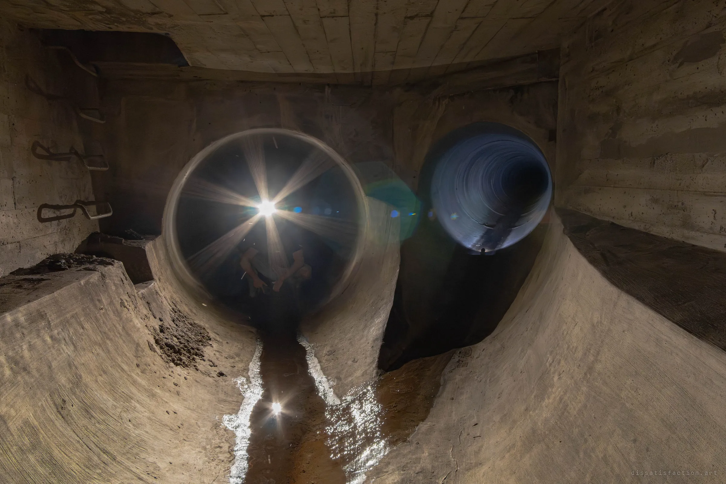 Inside a concrete junction chamber with steel ladder rungs, two pipes split off: in the left one an explorer crouches behind a bright starburst of light, and in the right one a second light glows far down a blue-lit tunnel. Water runs over the sandy floor below them.