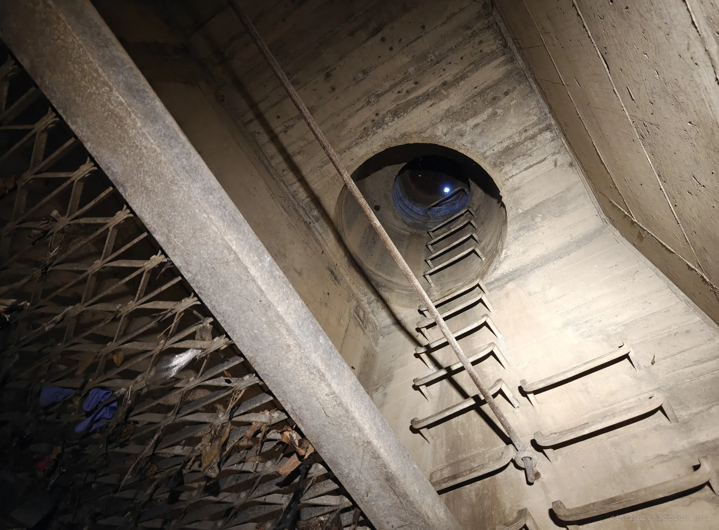 Looking up a concrete access shaft, steel rungs climb toward a round opening where a light shines far above, beside a diagonal beam and a debris-clogged grate holding leaves and discarded blue gloves. It shows the only way in and out of the drain.