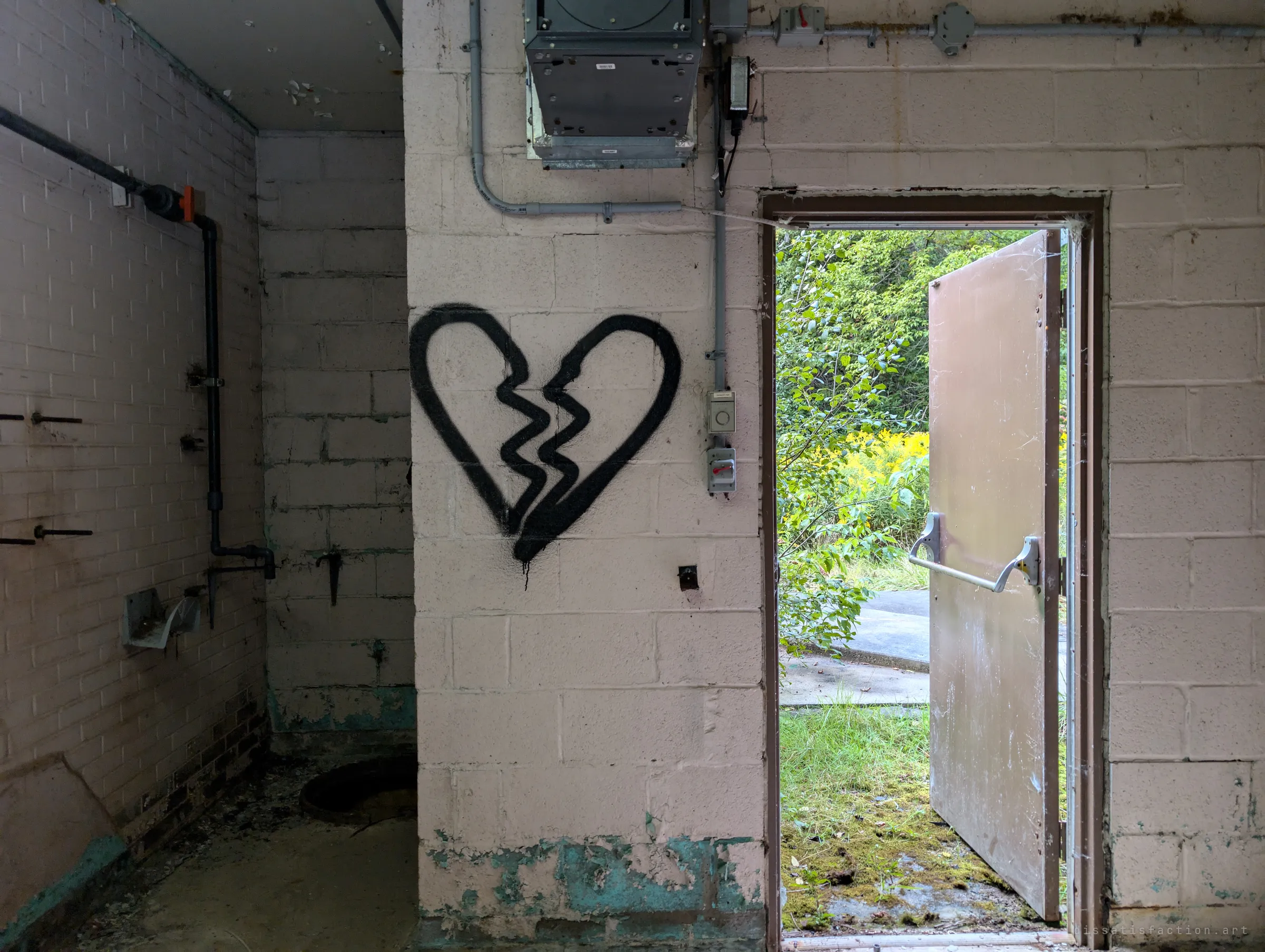 A broken-heart graffiti piece on a pale block wall beside an open steel door that lets in green overgrowth. Inside, a floor hatch and stripped pipework hint at the well below.
