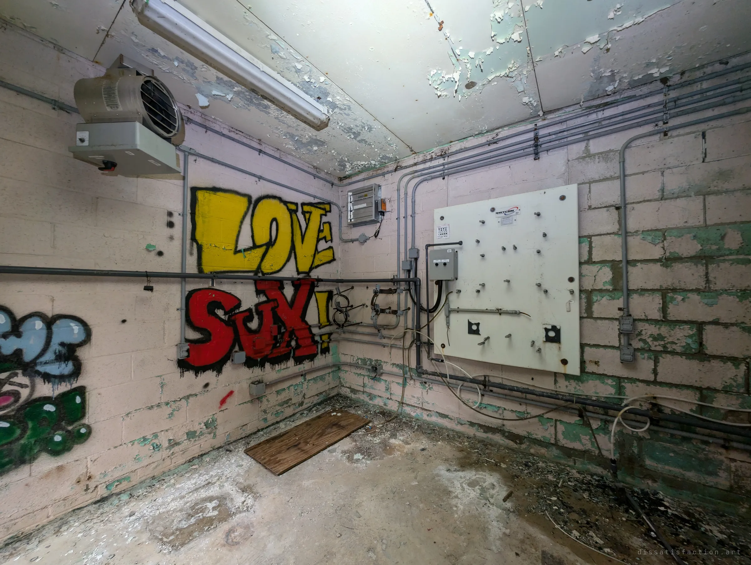 A corner room with peeling ceiling paint, a heater and a stripped white mounting board, the wall shouting 'love sux!' in yellow and red graffiti. It shows the control room gutted of its equipment.