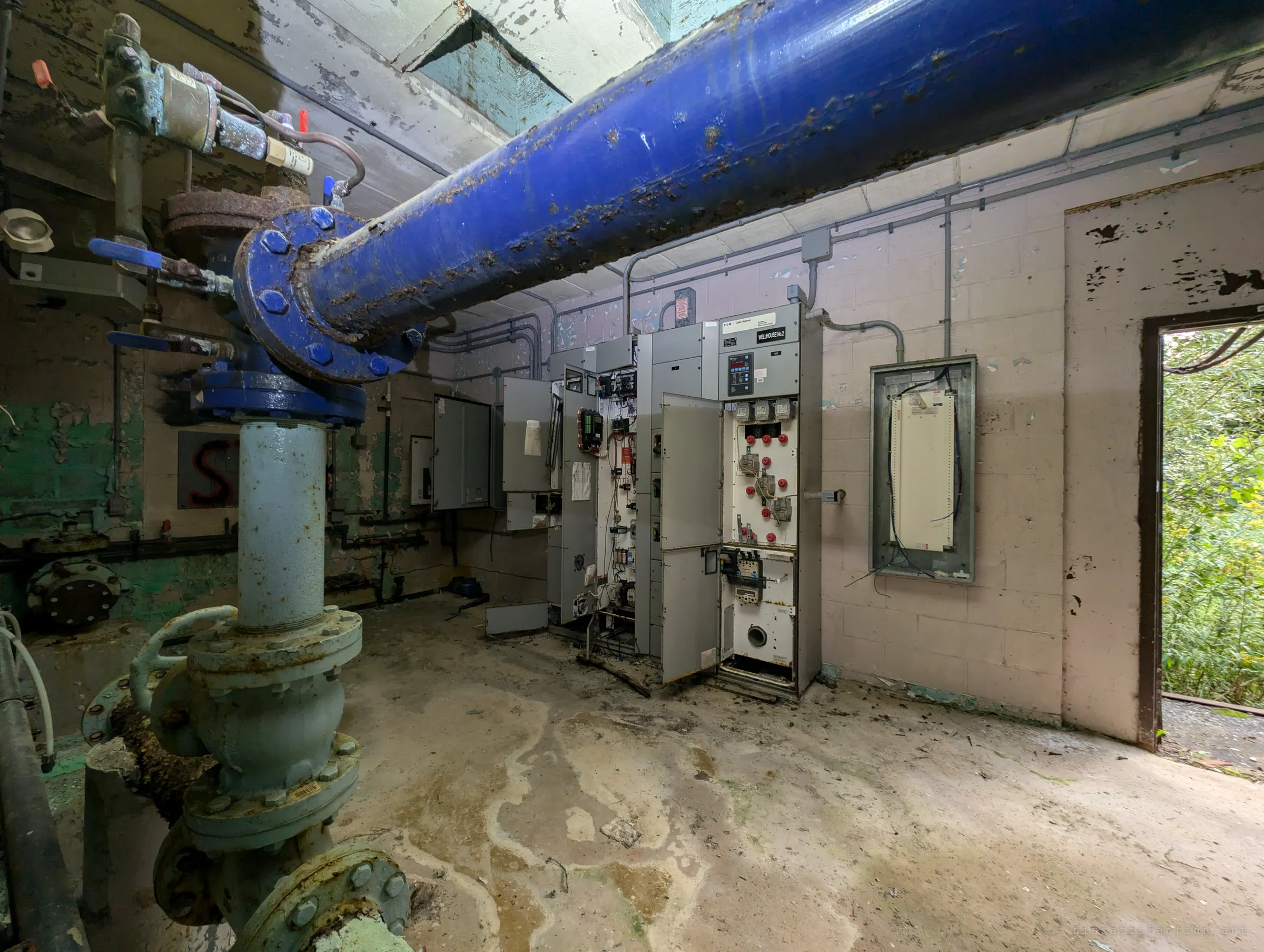 A fat blue water main runs overhead past a rusted valve stack toward a row of opened electrical cabinets. The open doorway on the right shows how the woods press right up to the pump room.