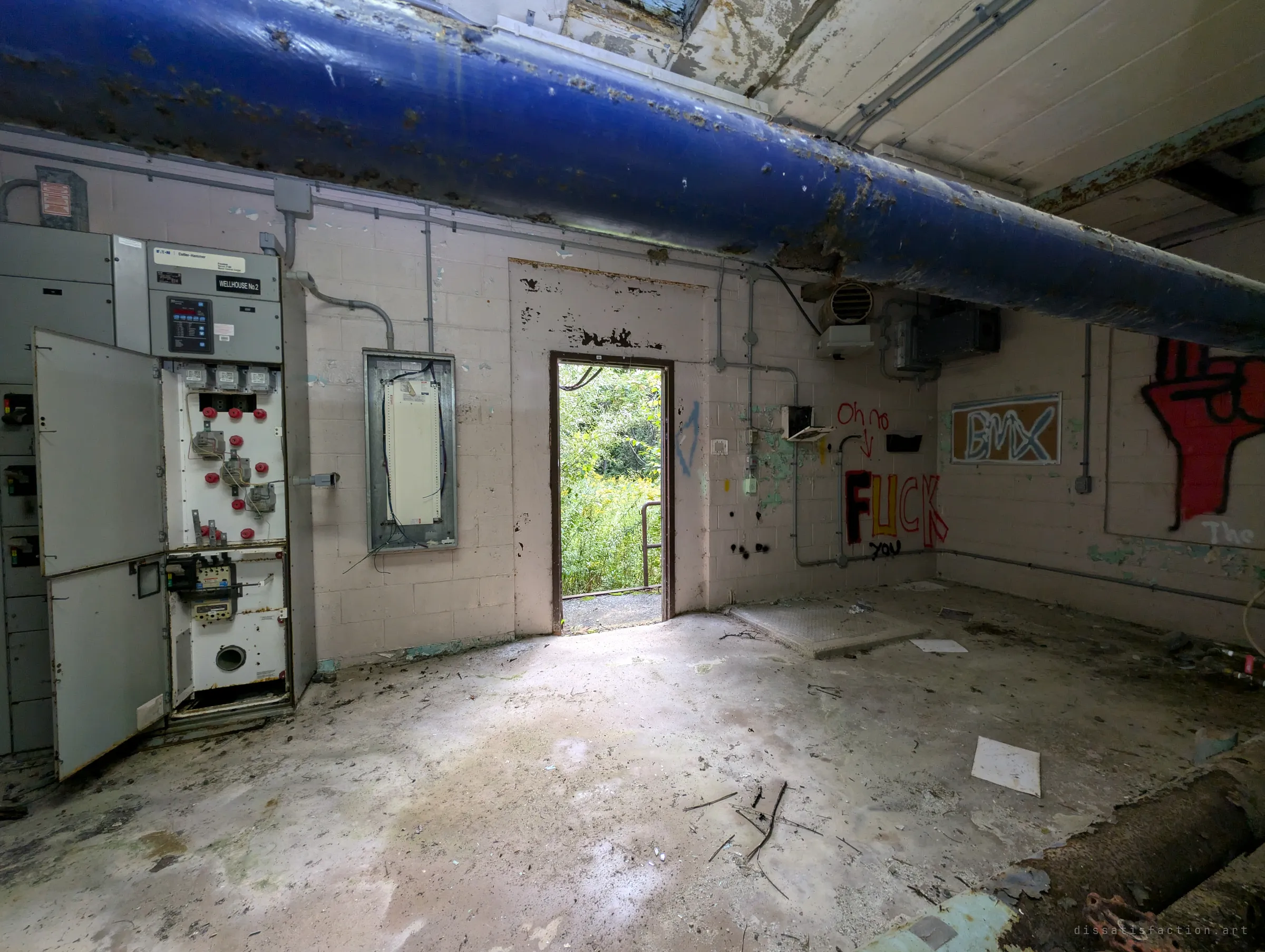The same pump room seen from the other side, with the blue main crossing the ceiling, gutted switchgear on the left and graffiti around a bright doorway. Litter and debris scatter the stained concrete floor.