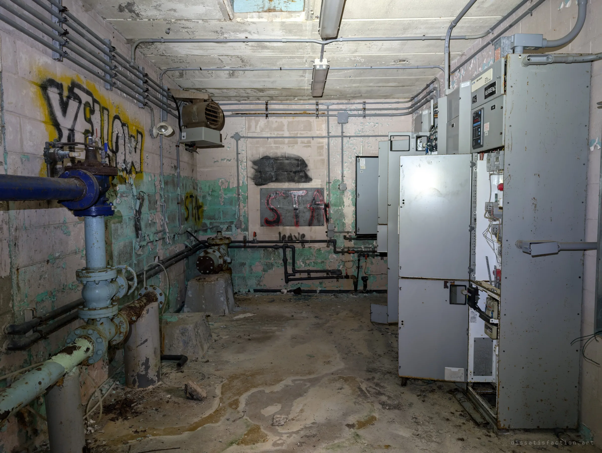 A narrow machinery aisle with pipework and a valve on the left and tall grey switchgear cabinets on the right, walls peeling green under tags. It captures the cramped layout of the well's pump hall.