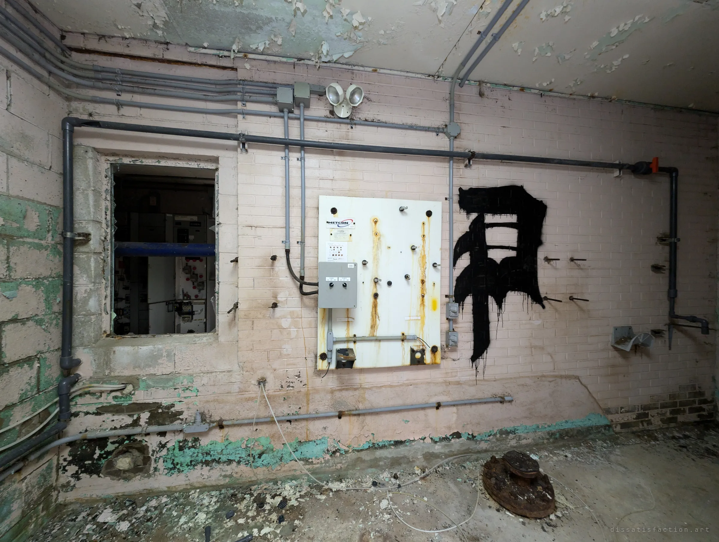 A rust-streaked mounting board and a bold black glyph painted on a pink brick wall, with an interior window looking back into the switchgear room. A capped pipe head sits on the debris-strewn floor.