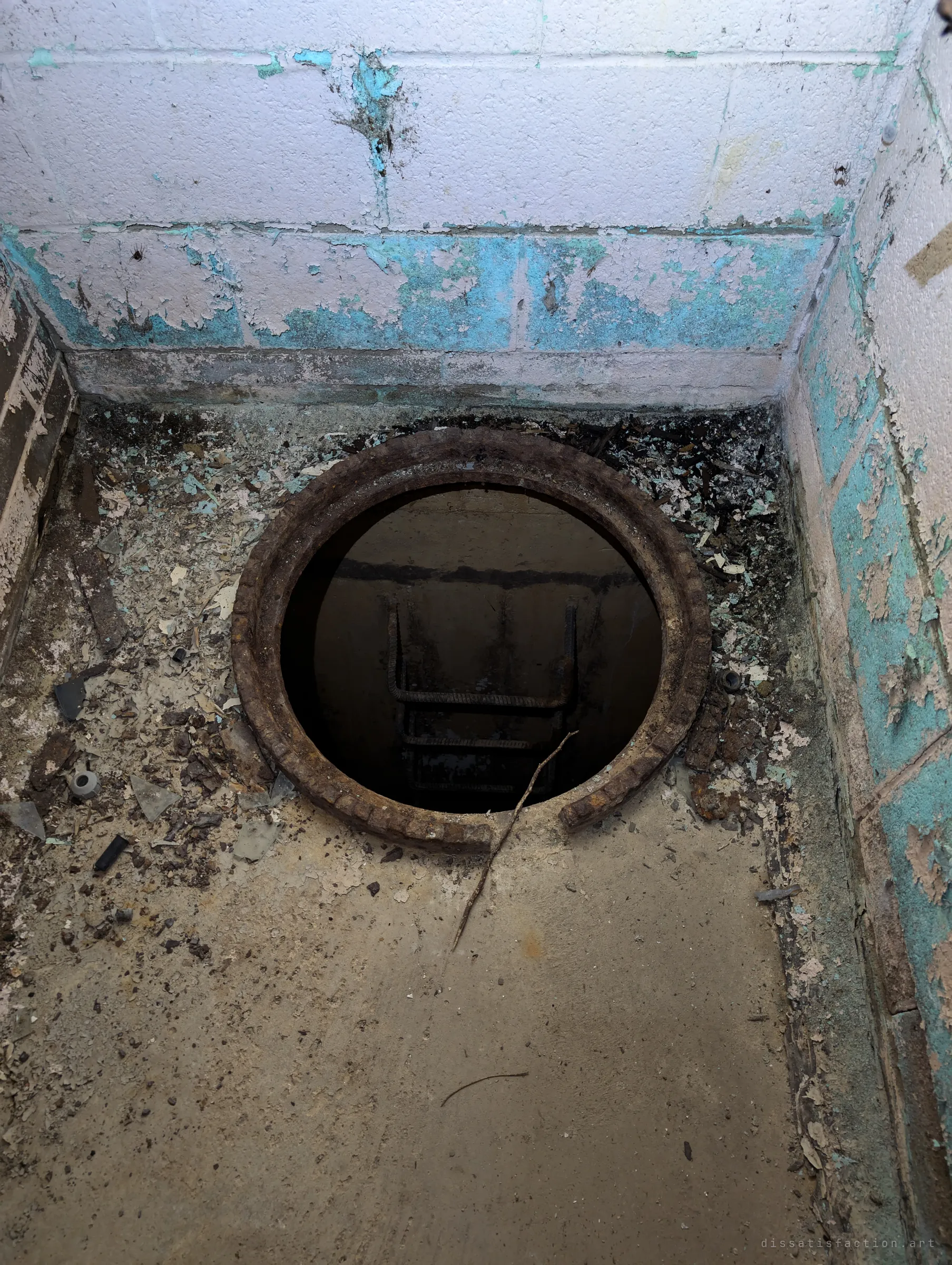 Looking down into a round hatch in a peeling turquoise-and-white alcove, with rebar rungs dropping into the dark shaft below. The opening leads down to the well's pipework.