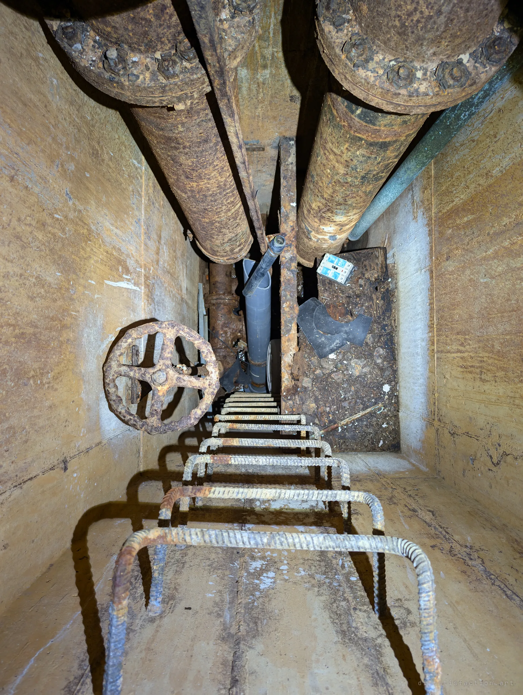 Straight down the shaft: rebar ladder rungs lead past a rusted valve wheel and two massive corroded pipes to the bottom of the well pit. The view shows the plumbing hidden beneath the pump house floor.