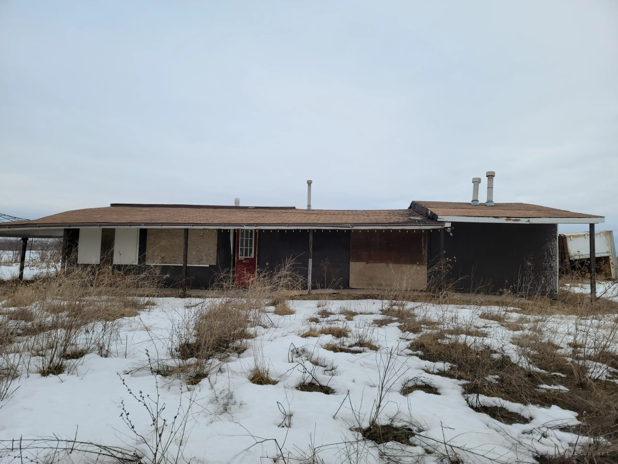 A long, low boarded-up building sits behind snow and dead grass under a flat grey sky, its red door the only colour left. The first look at the place, already emptied and sealed.