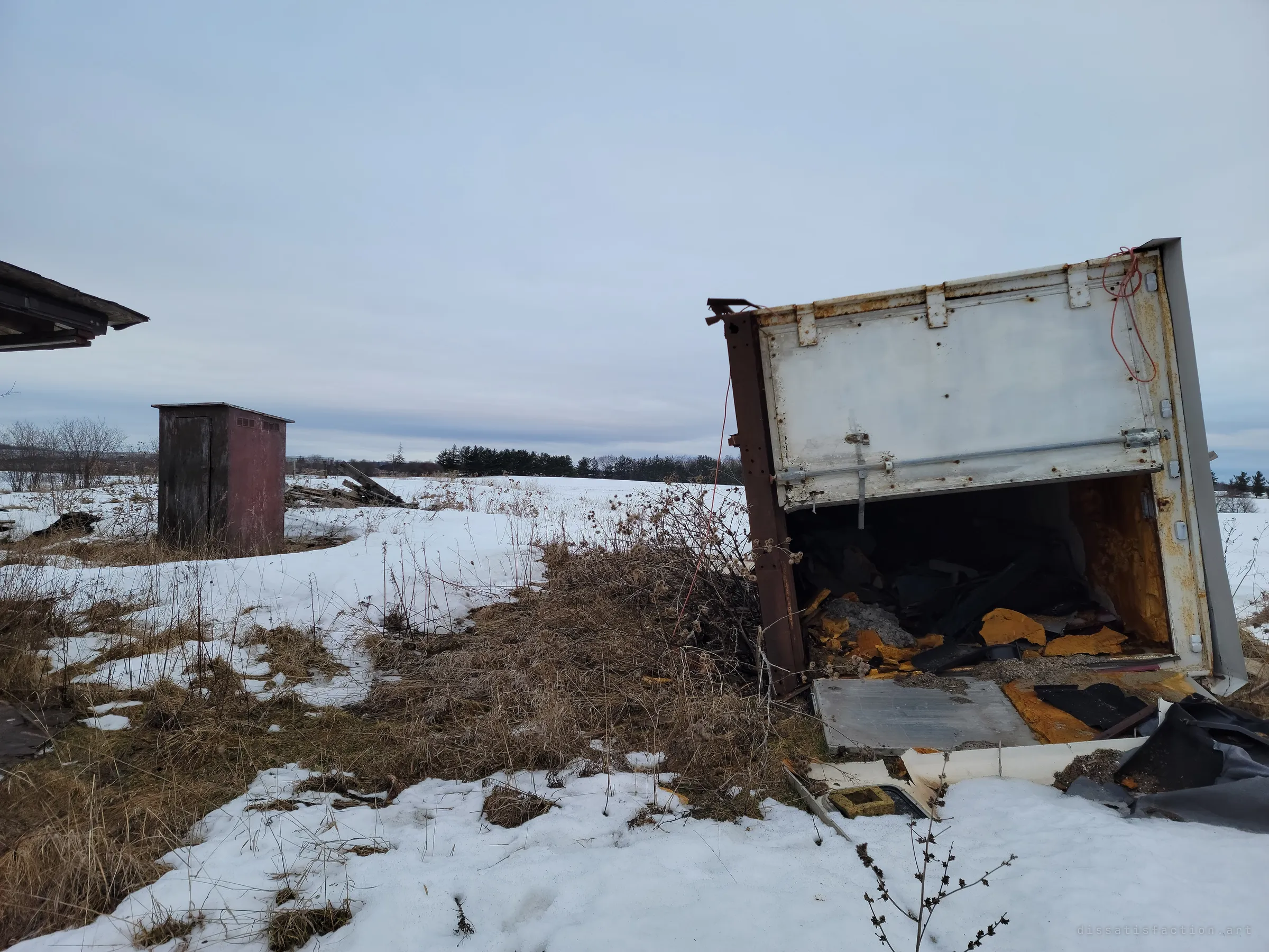 A tipped-over refrigerated truck box spills rusted foam into the snow beside a small weathered shed. Out here even the cold storage has been left to the weather.
