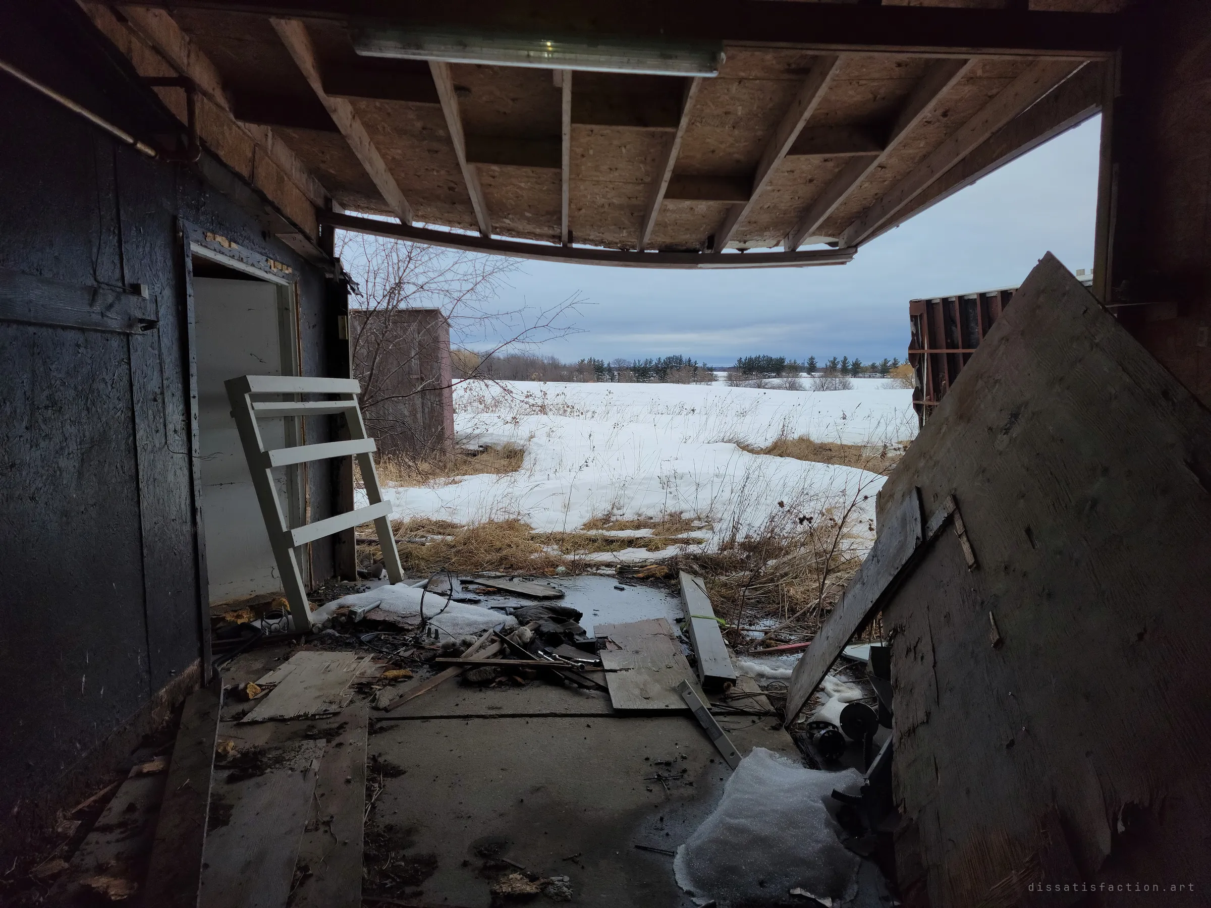 Looking out through a torn-open wall, a sagging ceiling frames a snowy field while broken boards and debris cover the floor. The building is opening itself to the landscape.