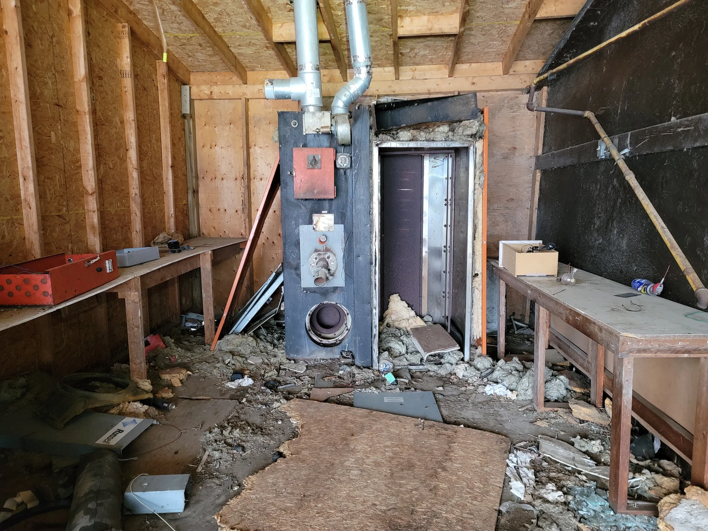 A tall black furnace with an open firebox door stands in a bare plywood room, insulation strewn across the floor between two workbenches. The machinery that once kept the place warm, gutted and cold.