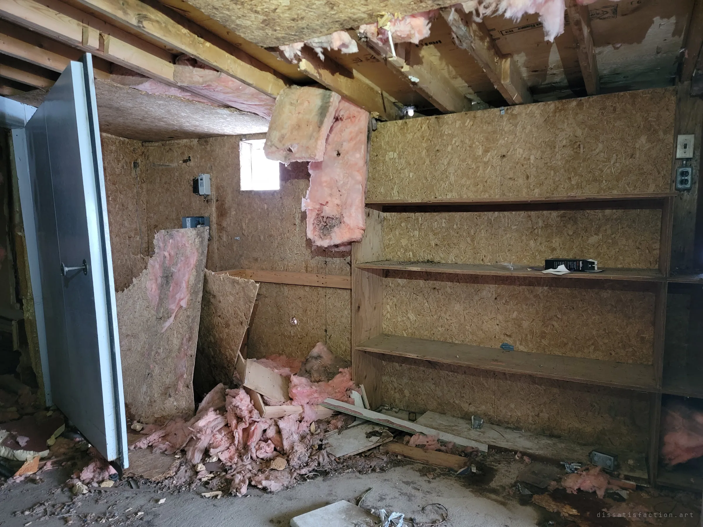 Pink insulation hangs from a collapsing ceiling over empty chipboard shelves, a heavy insulated door swung open at the left. The back rooms are coming apart from the top down.