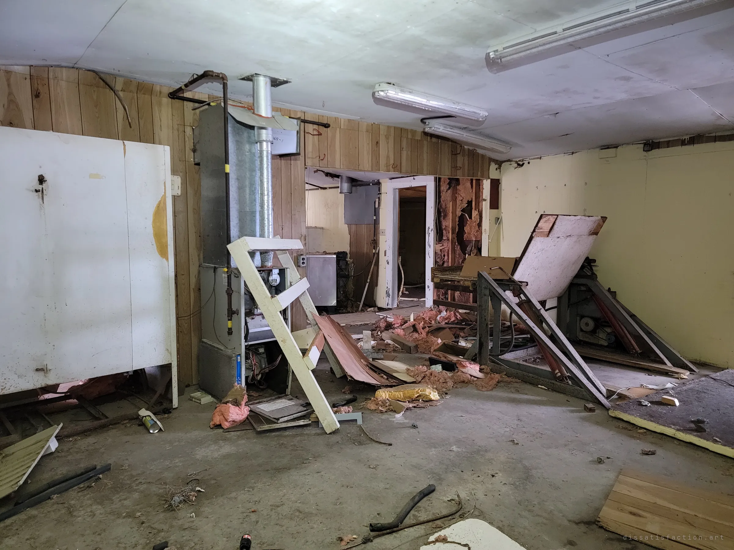 A wide workroom with wood-panelled walls, a stripped furnace, a fallen ladder and toppled equipment under dead fluorescent tubes. Everything useful has been pulled apart and dropped where it stood.