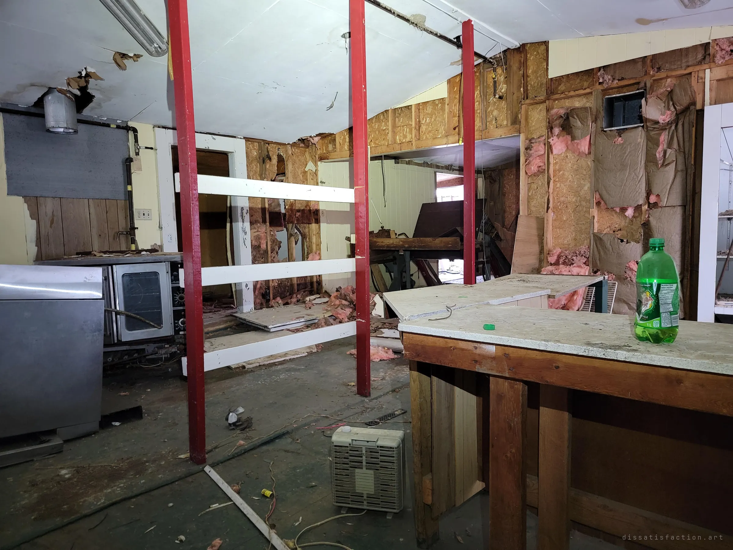 Red posts and white rails divide a gutted kitchen, an old convection oven on the left and a soda bottle left on a bare counter on the right. Someone's last drink, still standing.