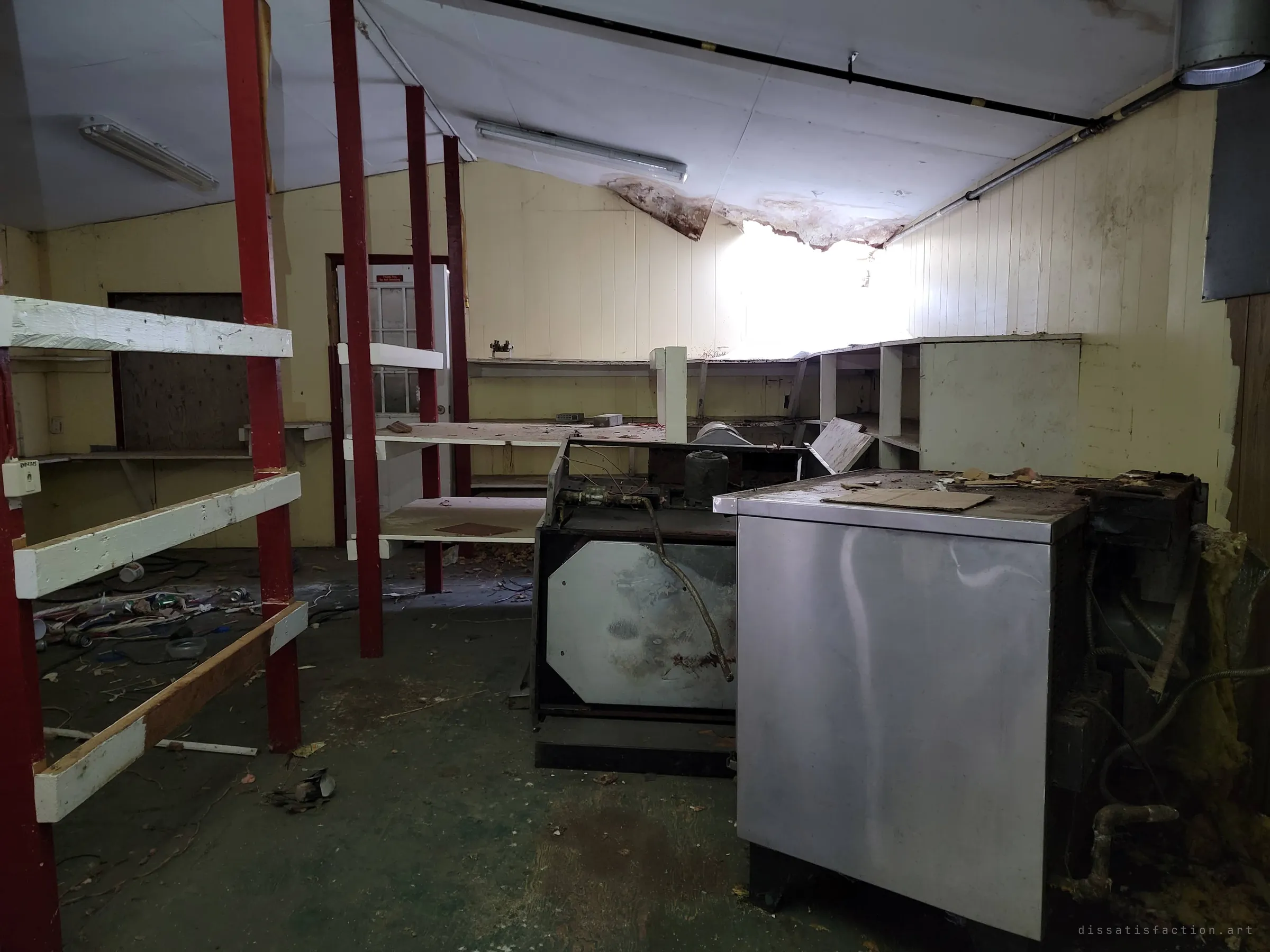 Stainless steel units stand abandoned in the middle of the shop floor, red-posted shelving to the left and a bright hole in the ceiling beyond. The counters are empty but the equipment never left.