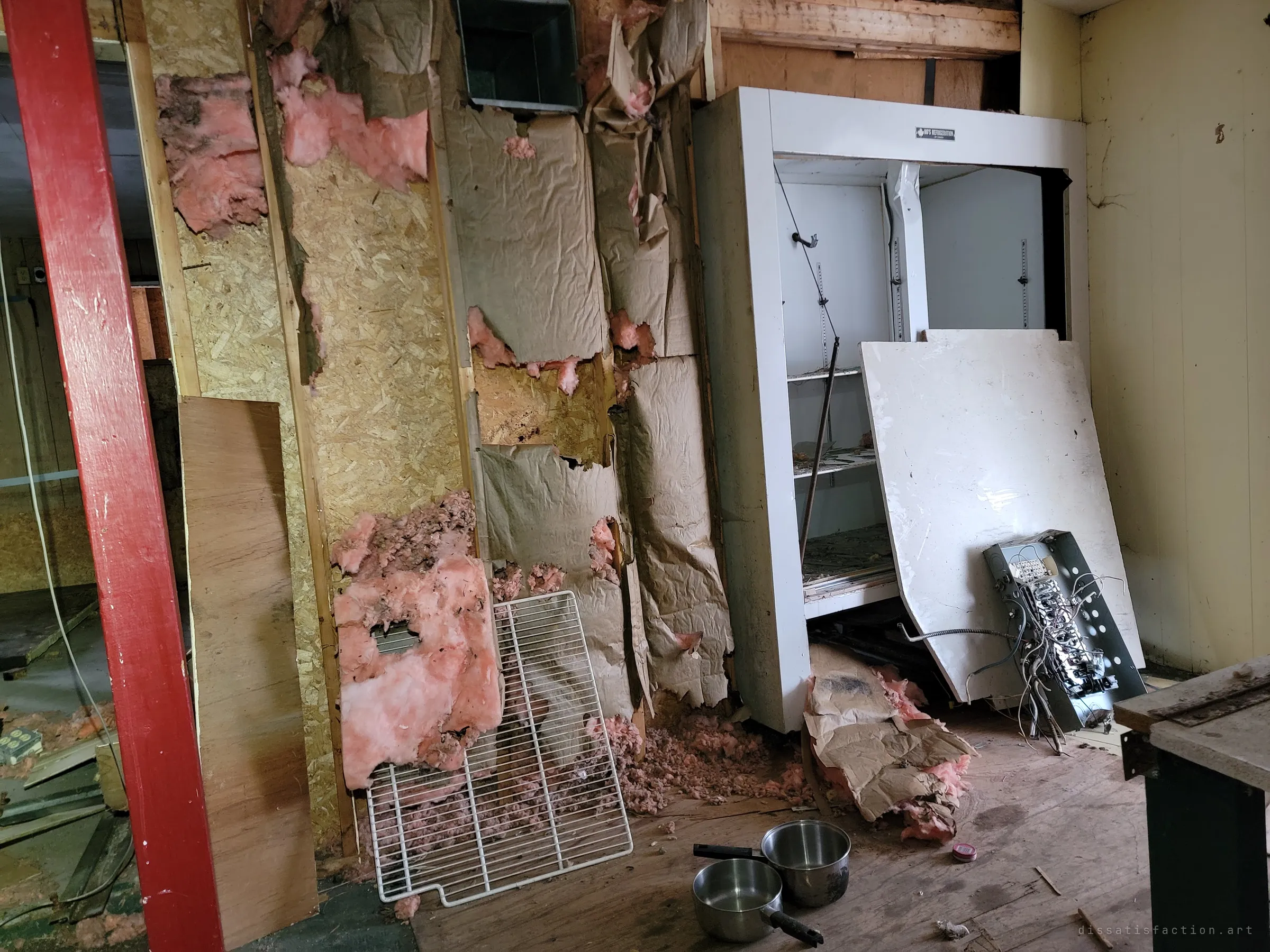 A doorless refrigerated display case sits against a wall stripped to torn paper and pink insulation, a pulled-out breaker panel and two saucepans on the floor. The kitchen reduced to its wiring and bones.