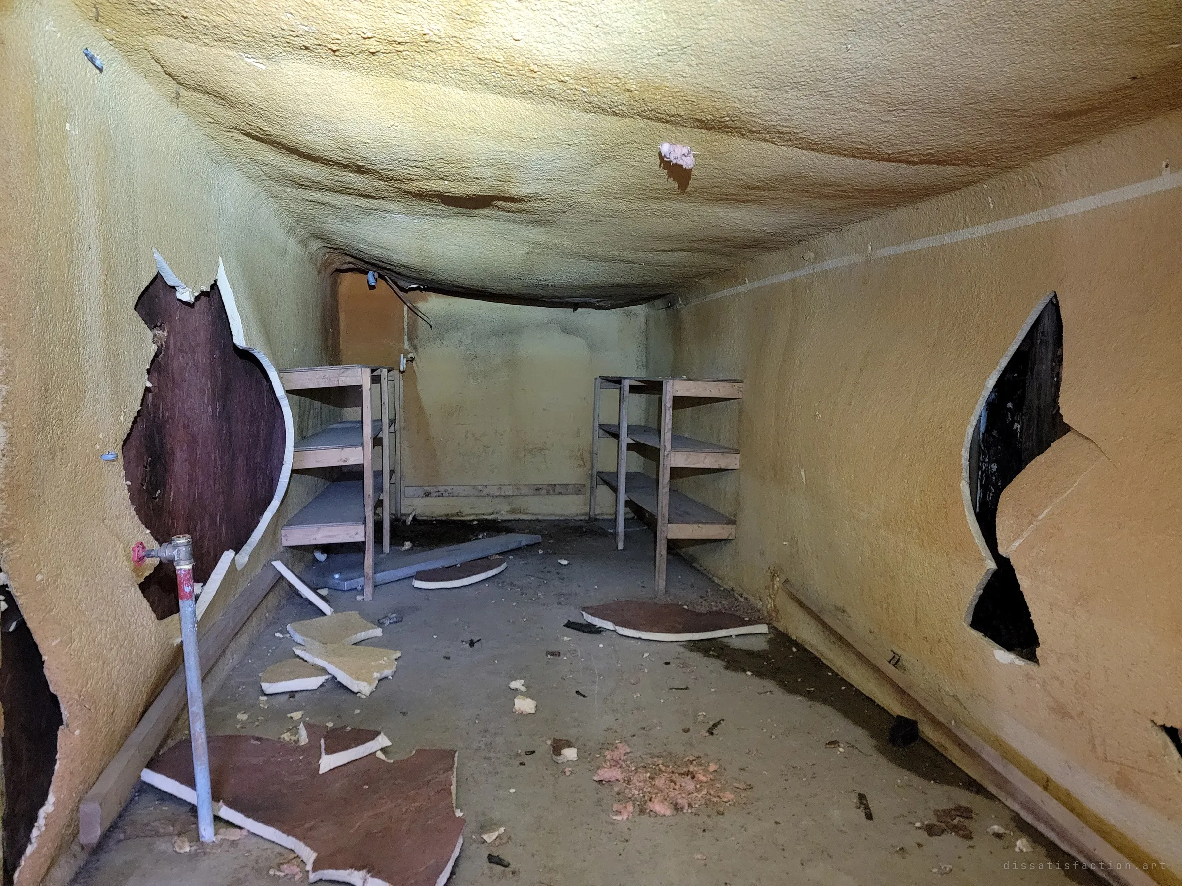 Inside a walk-in cooler, yellow spray foam sags from the ceiling and jagged holes are torn in the walls around two empty shelving units. A cold room that no longer holds anything, not even the cold.