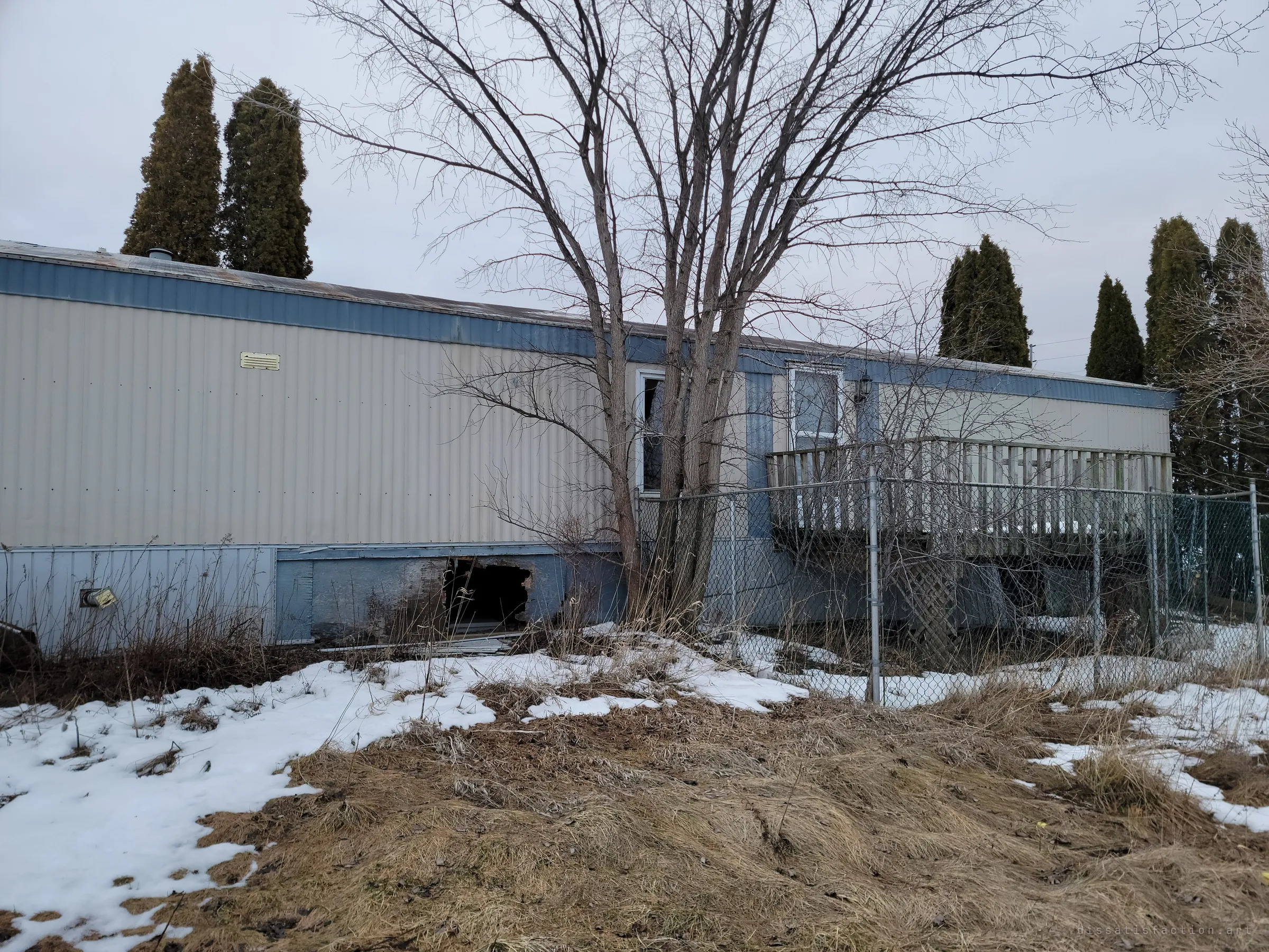 The long side of a beige and blue mobile home, its skirting torn open near the ground and a chain-link fence around the raised deck. The hole in the skirting shows how far the home has decayed.