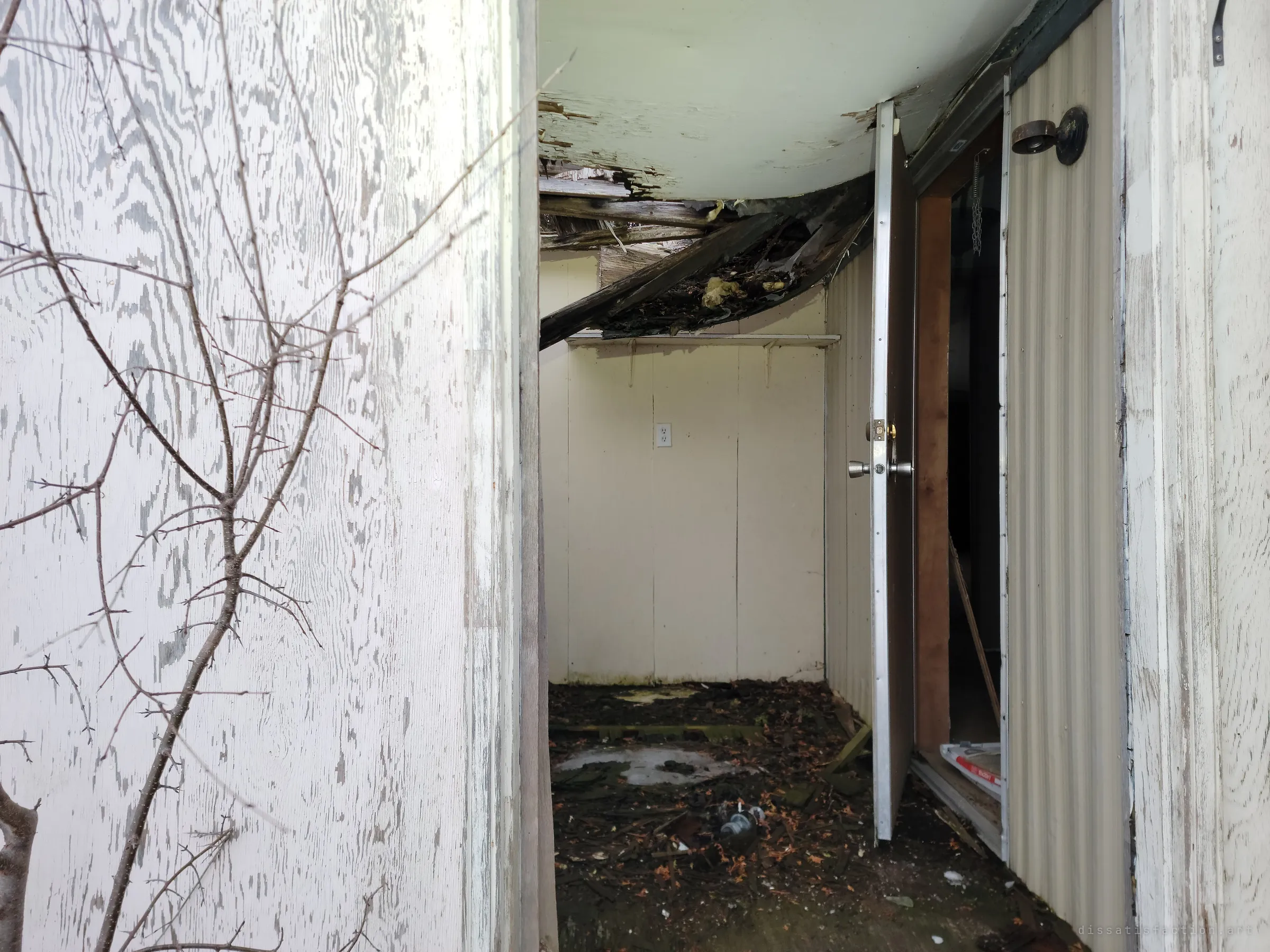 A lean-to entry porch with a collapsed, rotting ceiling sagging over a debris-strewn floor, the inner door left ajar. The caved-in roof shows water has been getting in for years.