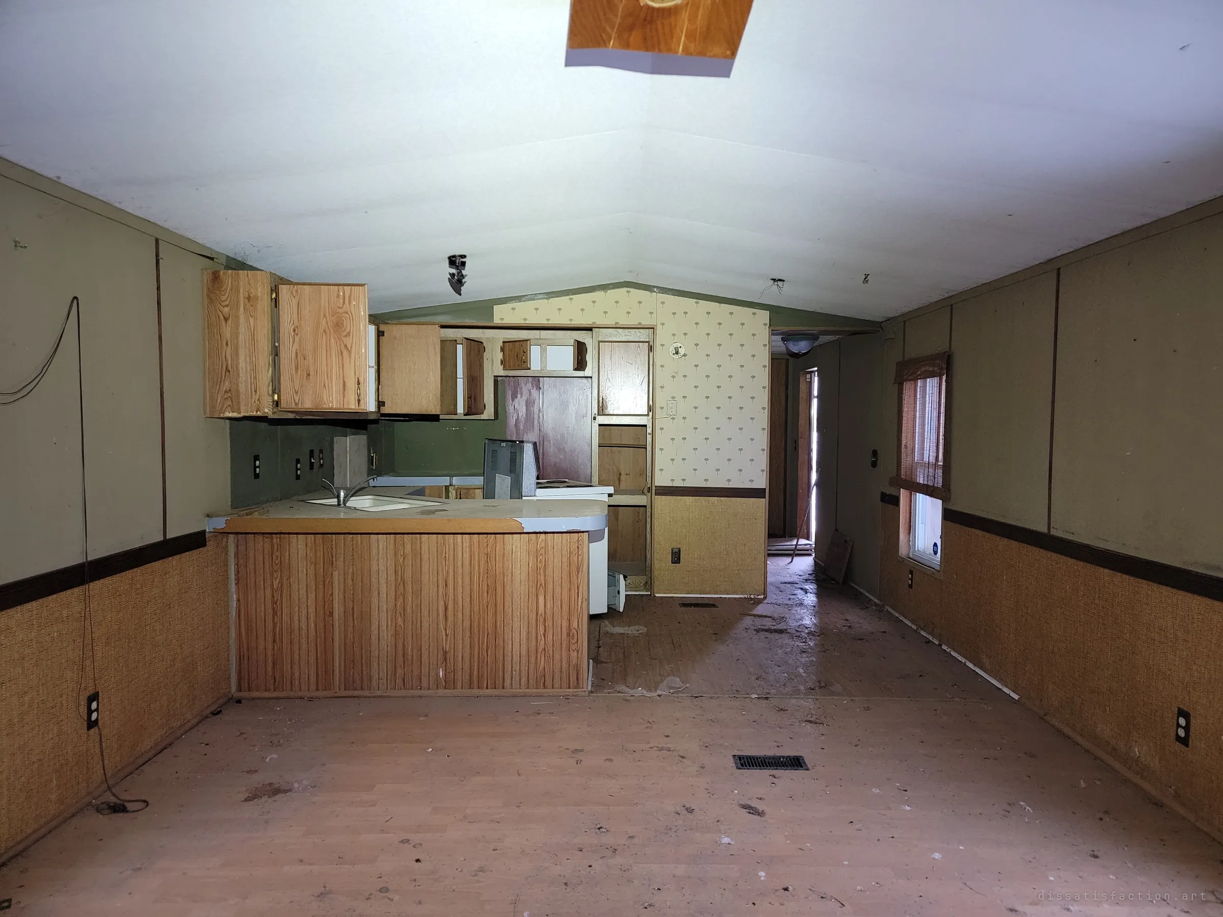 The open living room of the trailer, bare walls with wicker-textured wainscoting leading to a stripped kitchen and a dark hallway. A panel hangs loose from the ceiling above the empty floor.