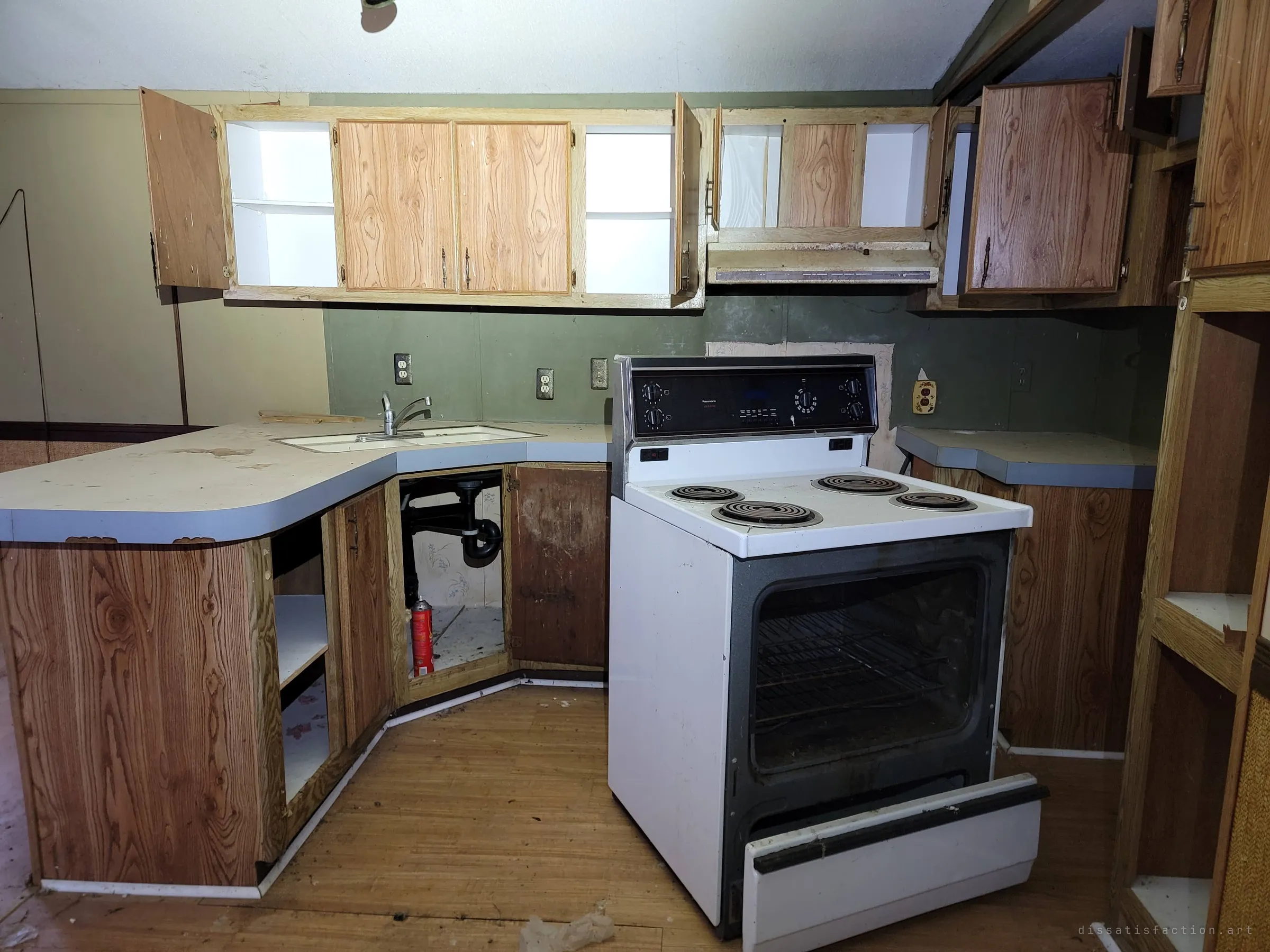 The kitchen with cabinet doors hanging open, an old electric range with its drawer pulled out and an empty cupboard under the sink. Everything useful is gone, but the fittings remain.