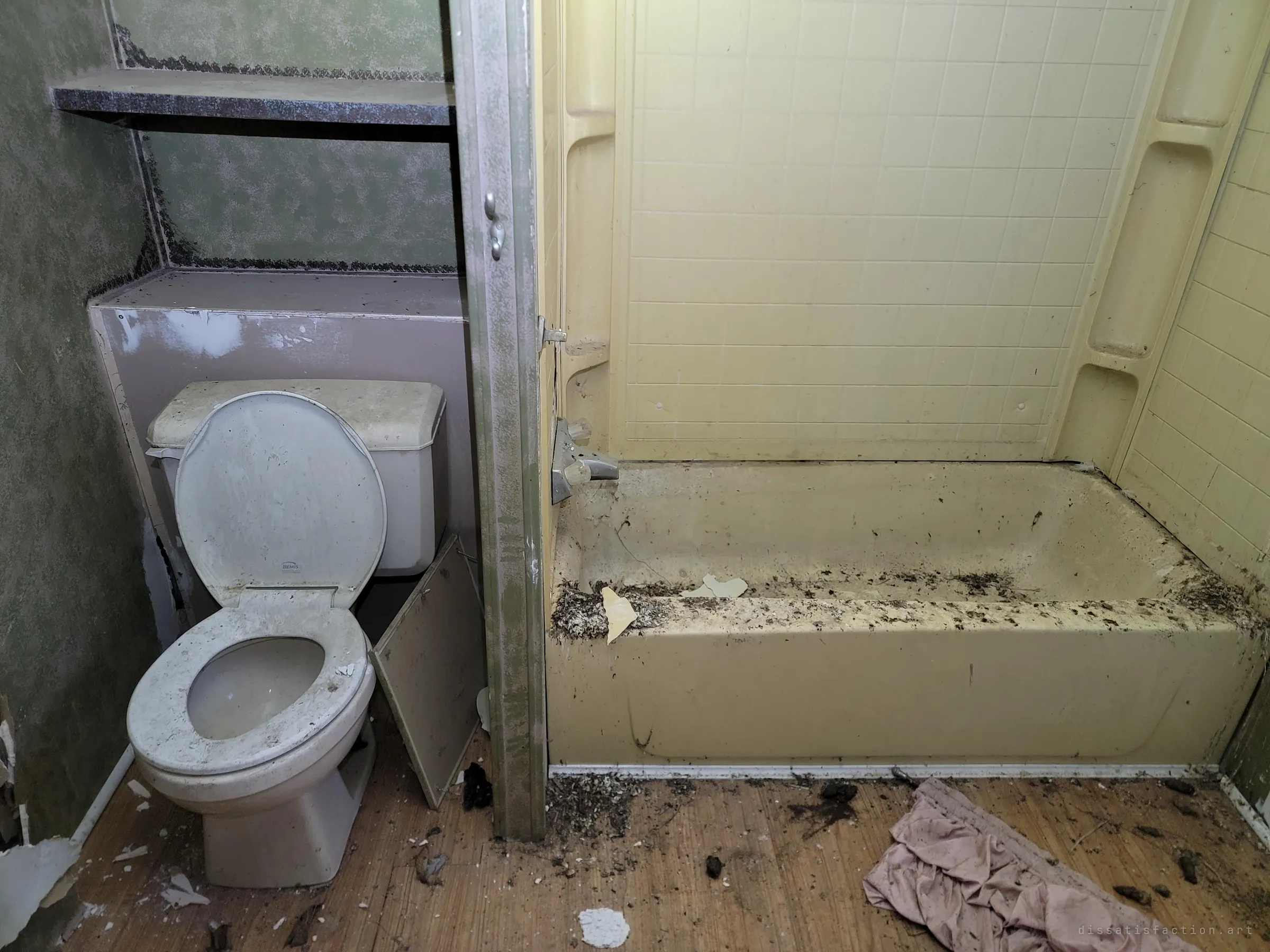 A bathroom with a grimy toilet and a yellow tub littered with droppings and debris, a pink cloth crumpled on the floor. Animals have clearly moved in since the people left.