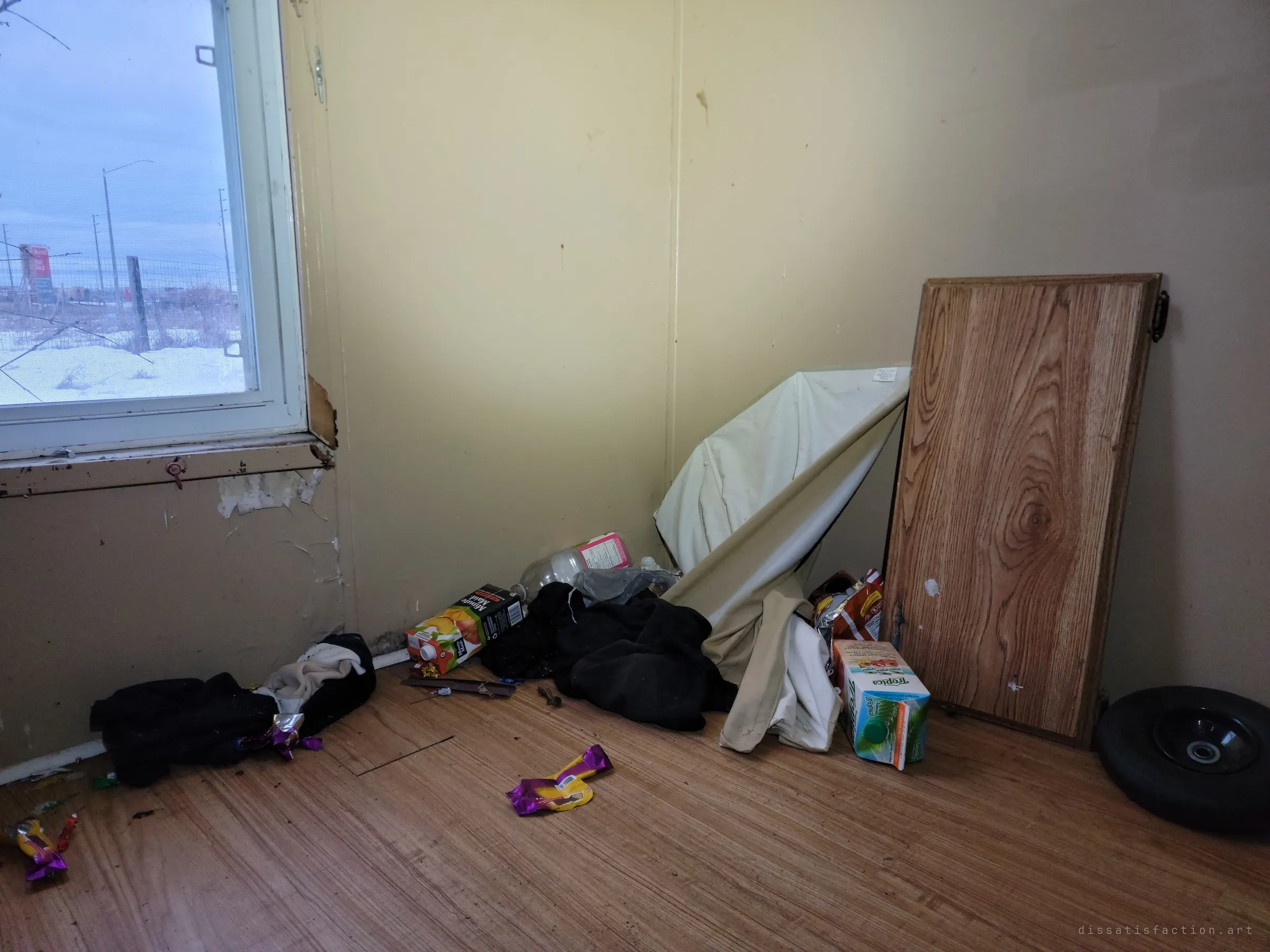 A bedroom corner piled with dropped clothes, juice cartons and candy wrappers beside a leaning cabinet door, grey winter light coming through the window. Signs of later visitors camping out.