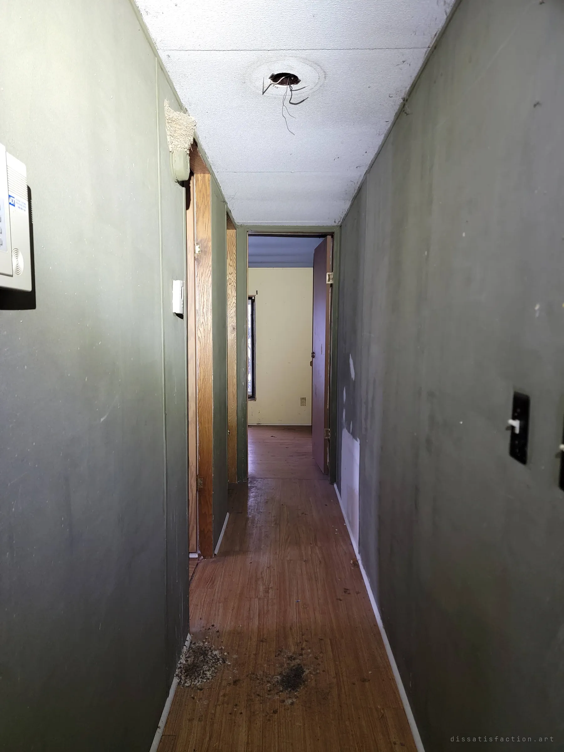 A narrow dark-green hallway running to a pale bedroom at the far end, an old alarm keypad on the wall and bare wires hanging from the ceiling where a light used to be.
