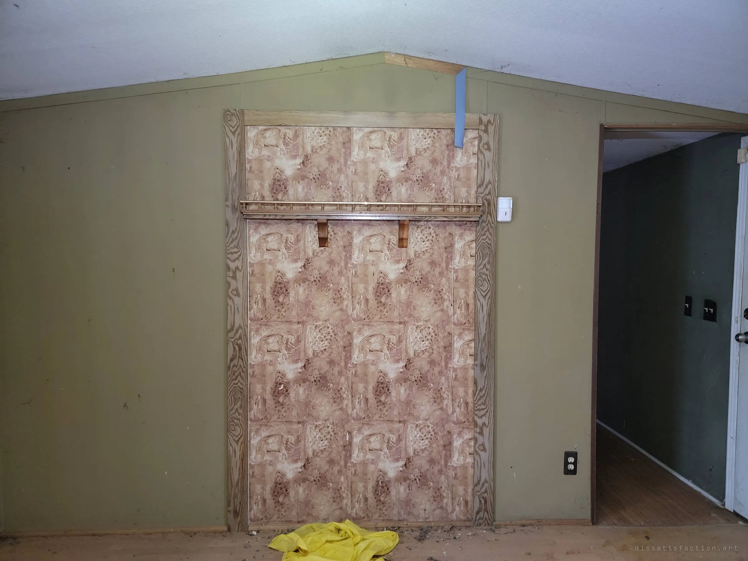 An elephant-print wallpaper panel framed in faux-grain trim, with a small shelf across it, set into an olive wall under the peaked ceiling. A yellow raincoat lies abandoned on the floor below.