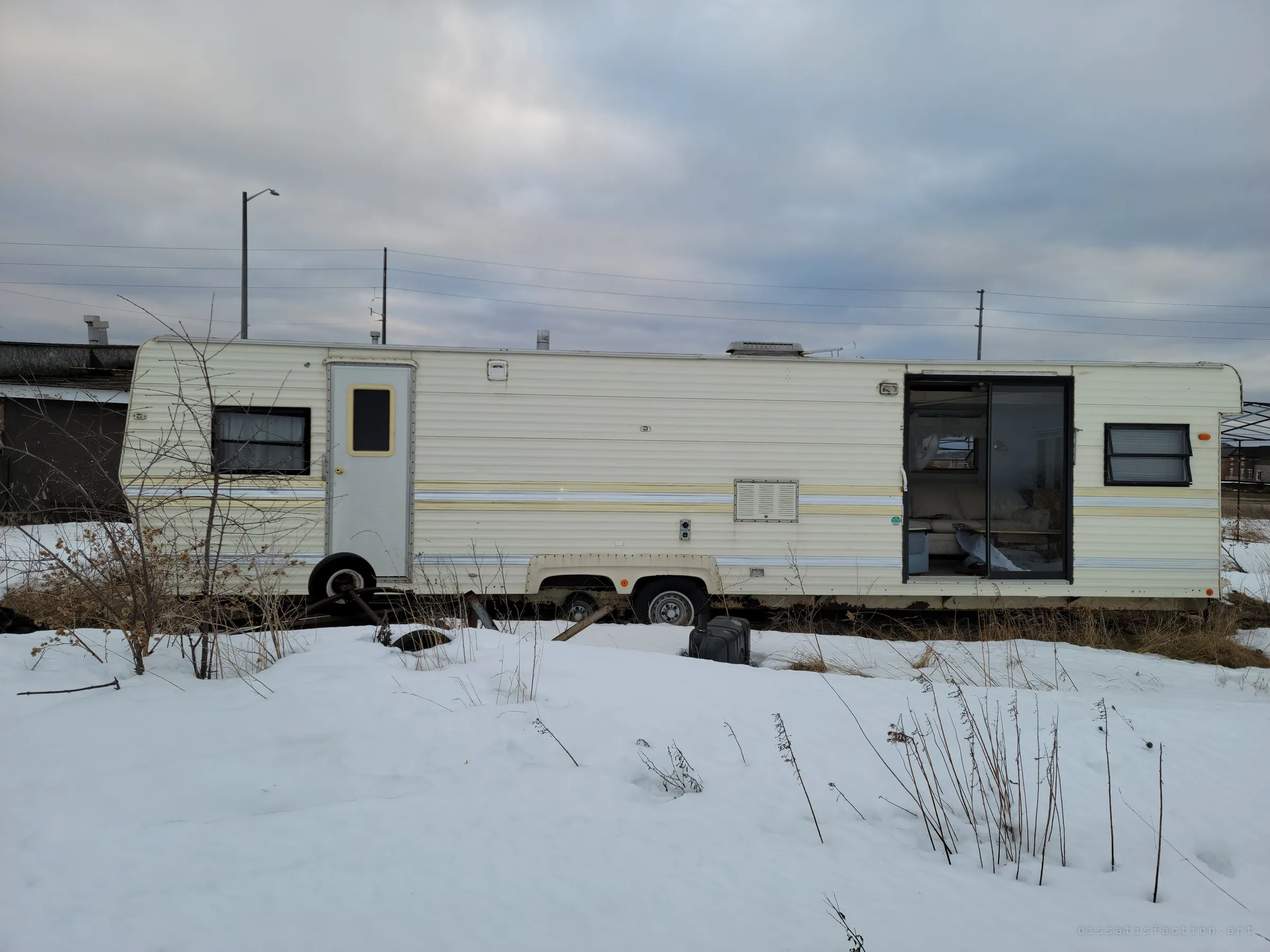 The full length of a cream travel trailer stranded in snow, its sliding door left open. Weeds and scrap sit around it, showing how long it has been parked for good.