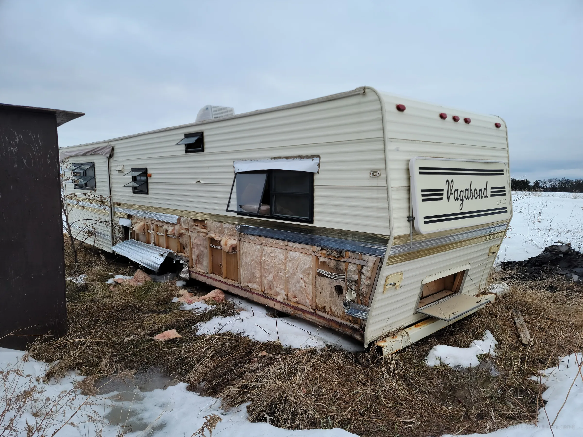 The back corner of the trailer, its lower siding torn away to expose pink insulation and rotting framing. The maker's badge still hangs on the end wall above the dead grass.