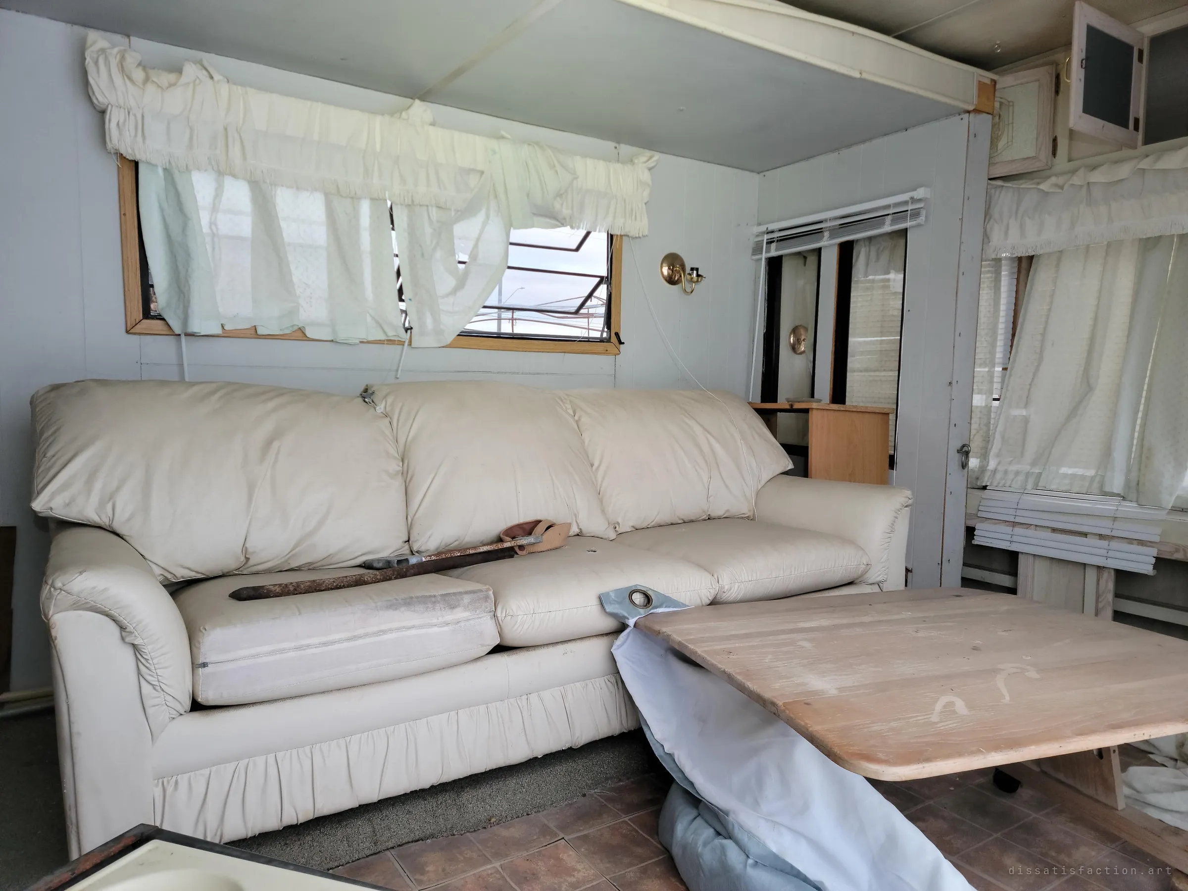 A cream leather sofa fills the trailer's living area under torn curtains, a rusty bar lying across its cushions. A collapsed table leans into the frame.