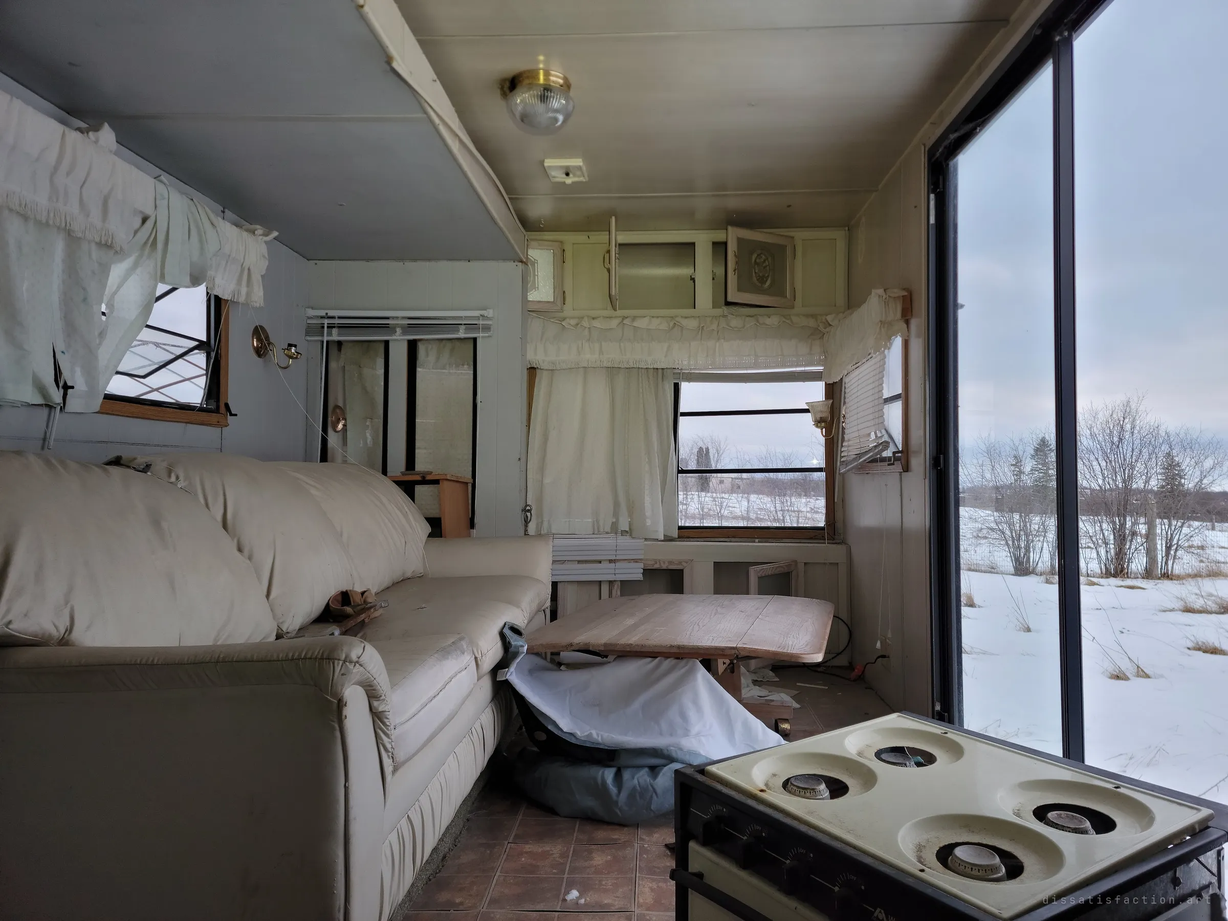 Looking down the trailer from the stove toward the front windows, the sofa and a sagging table in between. Winter light pours through the open sliding door.