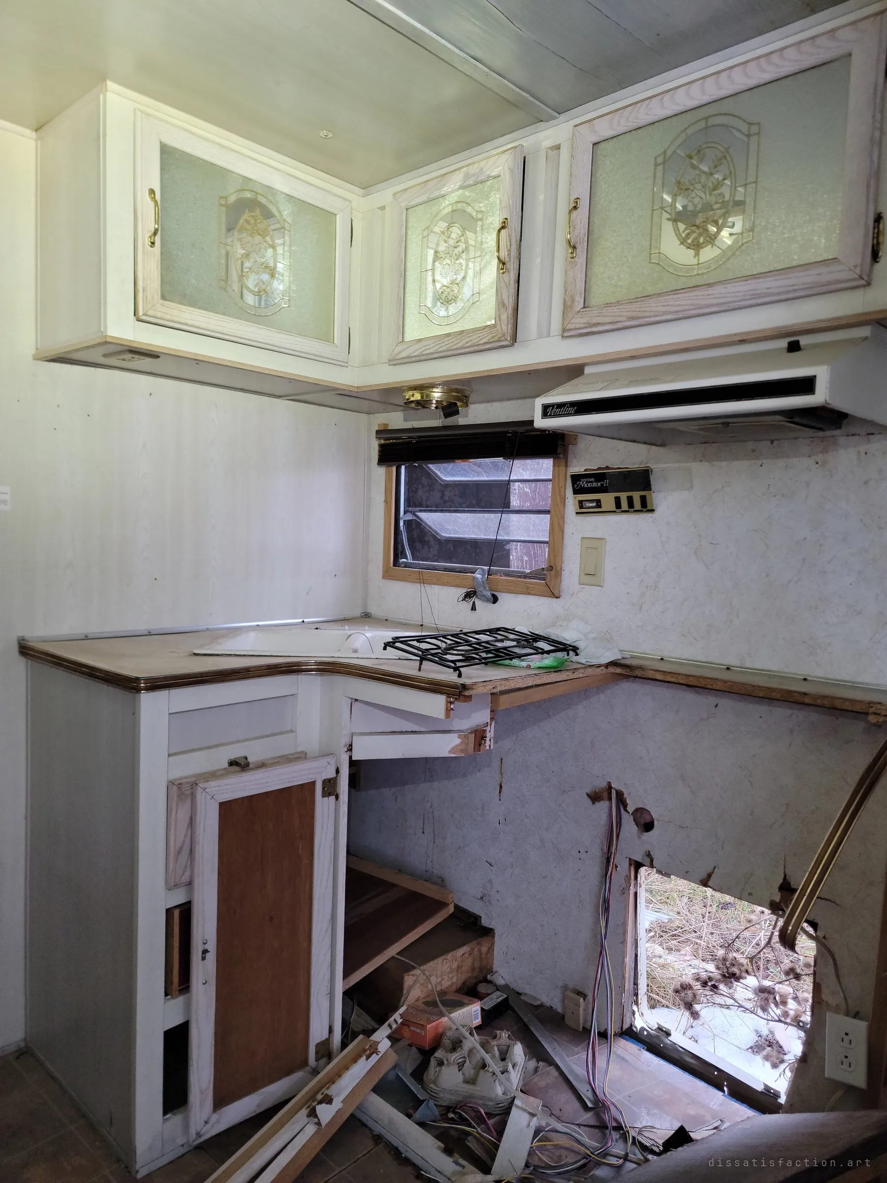The corner kitchen, with frosted cabinet doors still in place over a gutted counter. Below, the wall has been broken through to the dead grass outside, with loose wiring spilling out.