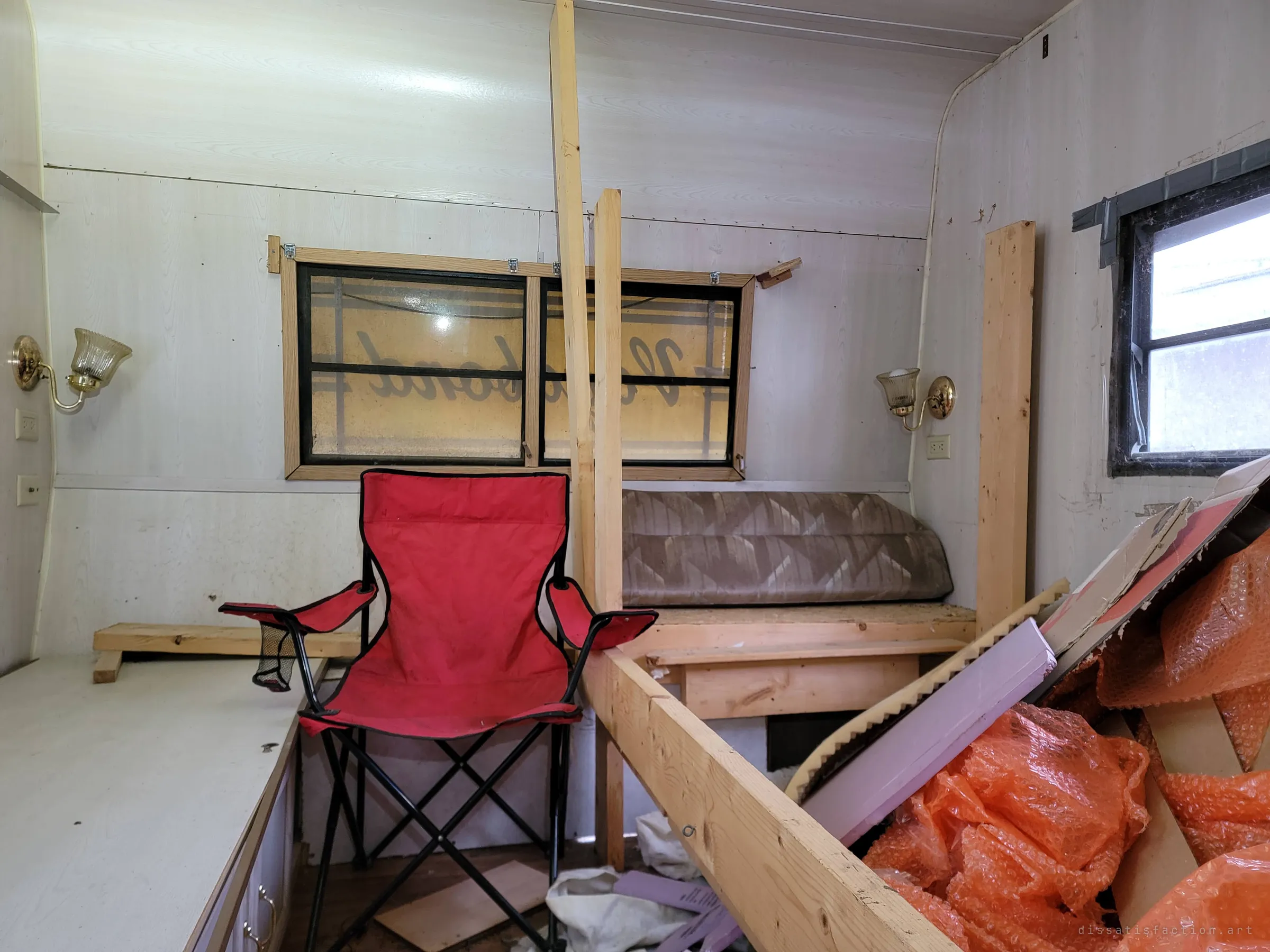 A red camp chair stands among lumber and orange bubble wrap in the bedroom. The trailer's own badge shows backwards through the window behind it.