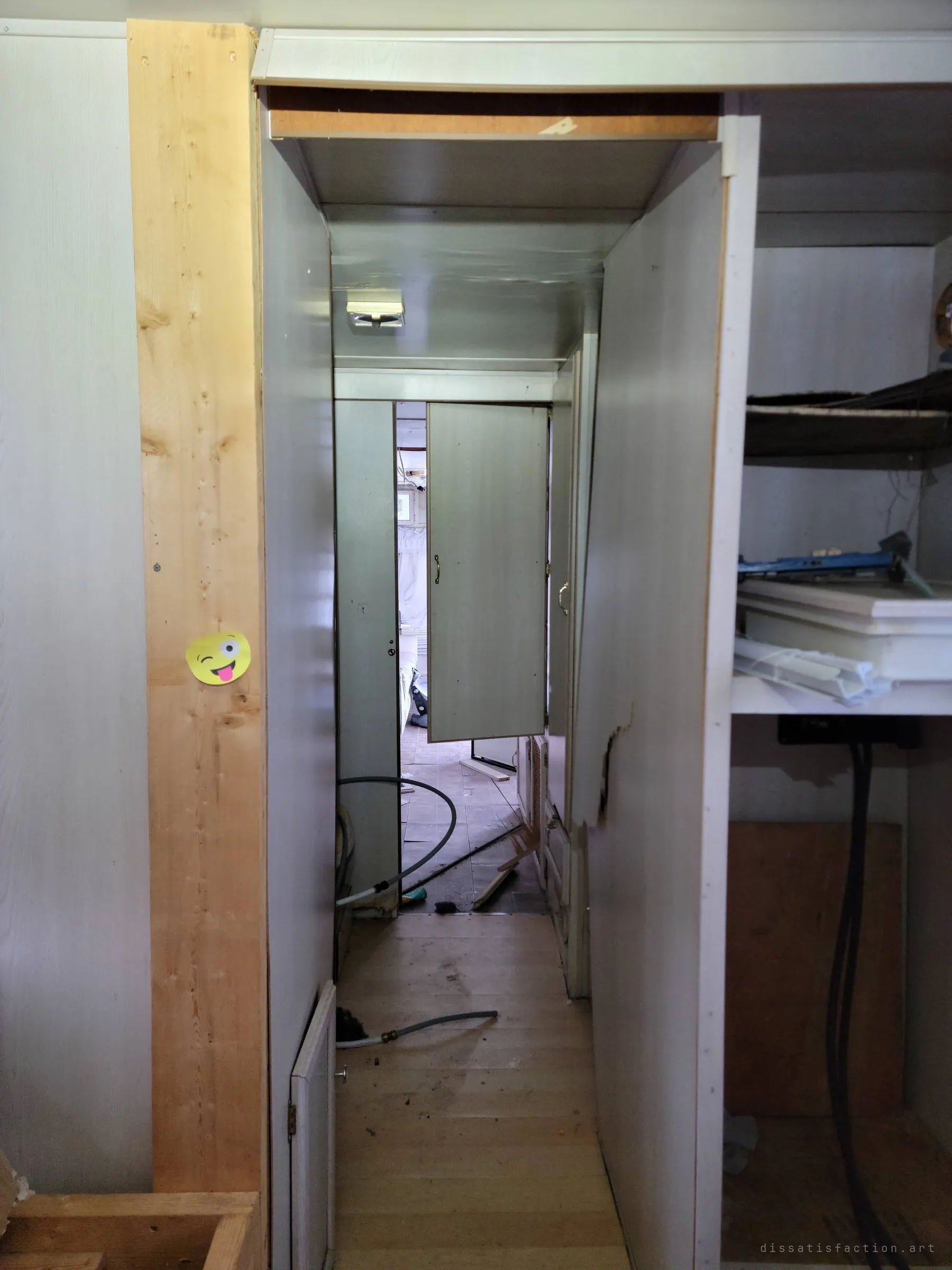A narrow hallway runs the length of the trailer past a door hanging open, a winking emoji sticker on the stud beside it. Hoses snake across the floor.