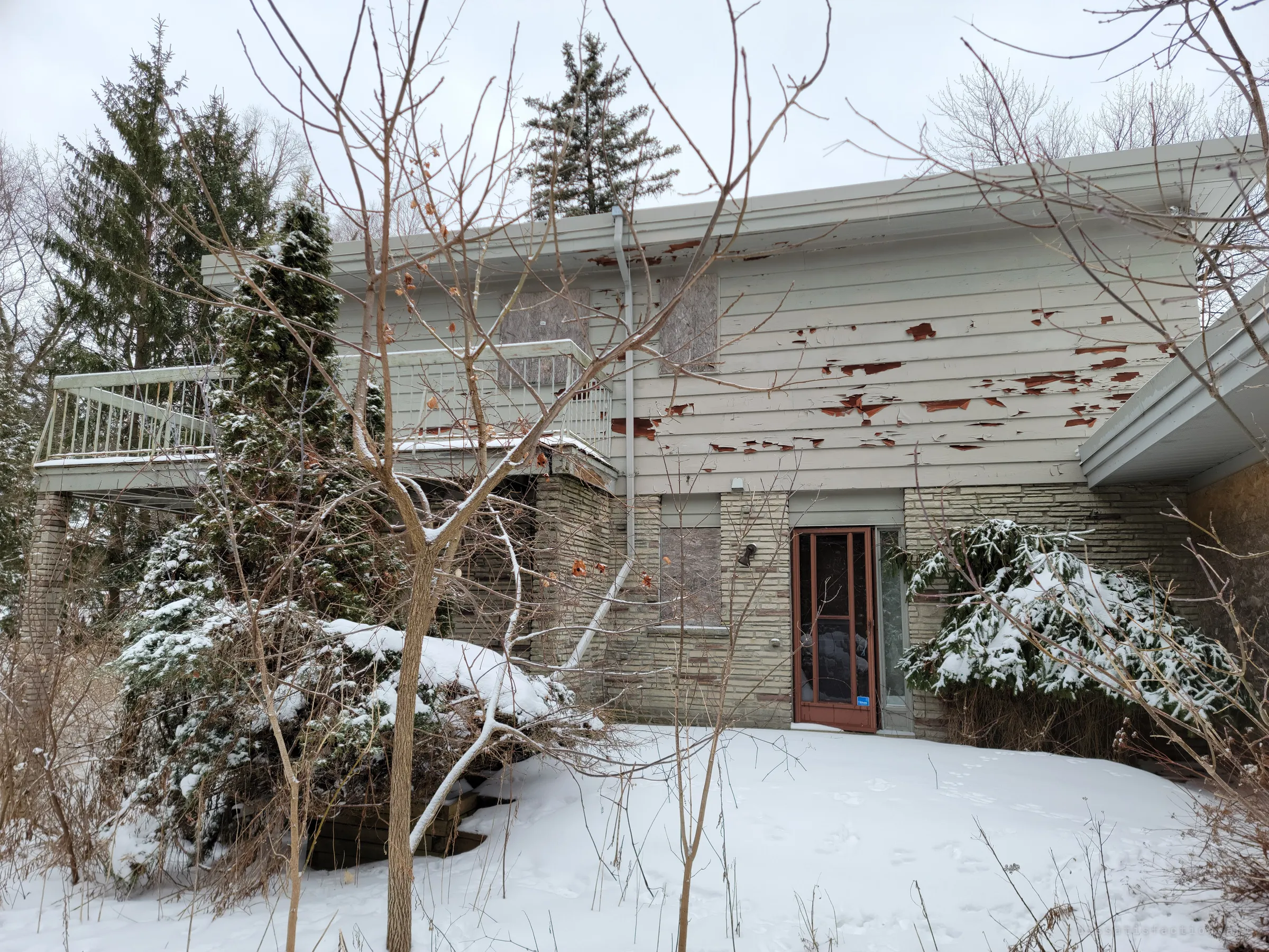 The back of a two-storey mid-century house behind snowy shrubs, its upper windows boarded and its siding peeling in rust-red patches. A wraparound balcony and a lone storm door hint at the home it used to be.