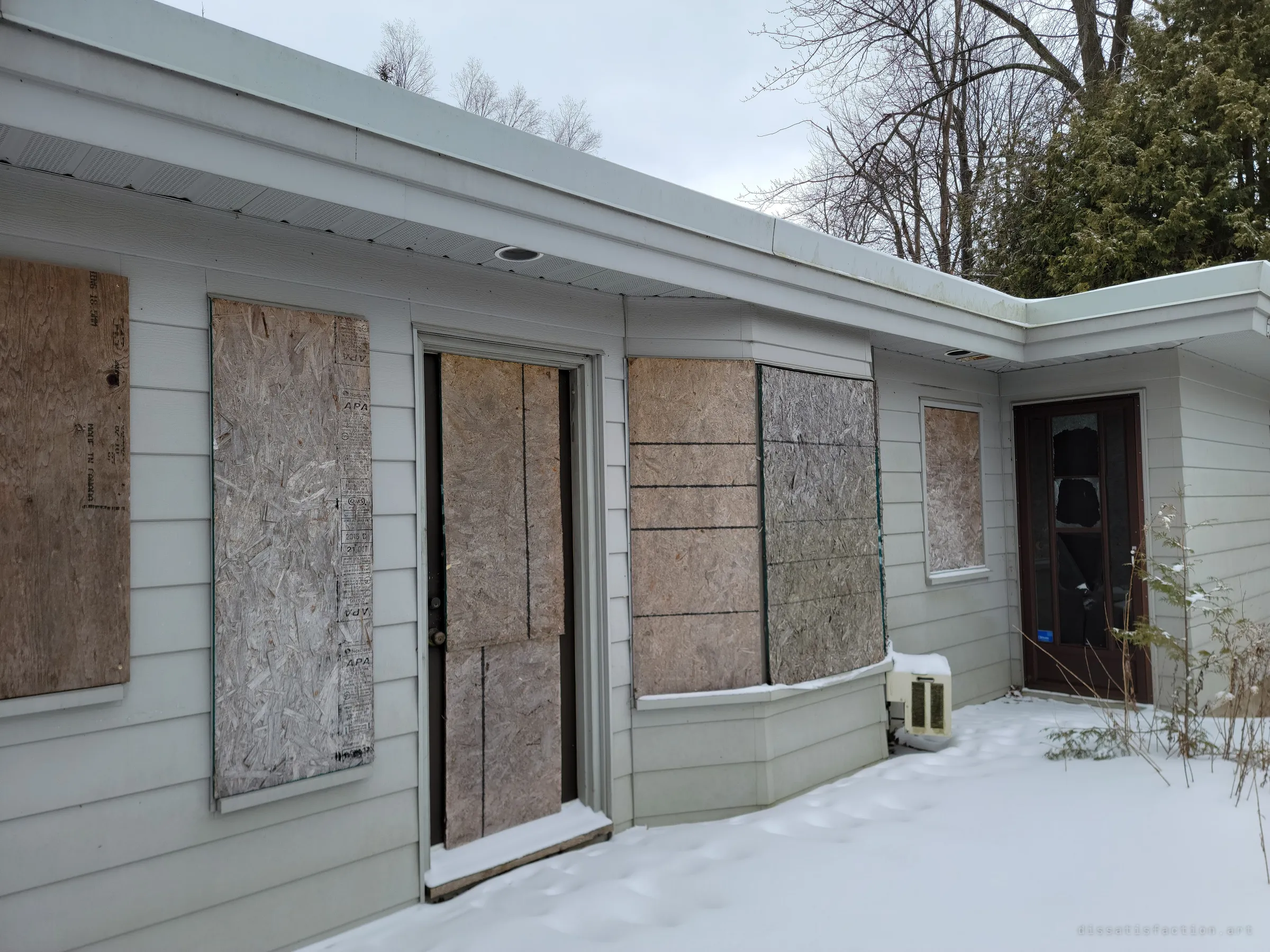 A long low wing of the house with every window and a door covered in plywood, and a glass door at the end already smashed. Sealed up, but not well enough to keep people out.