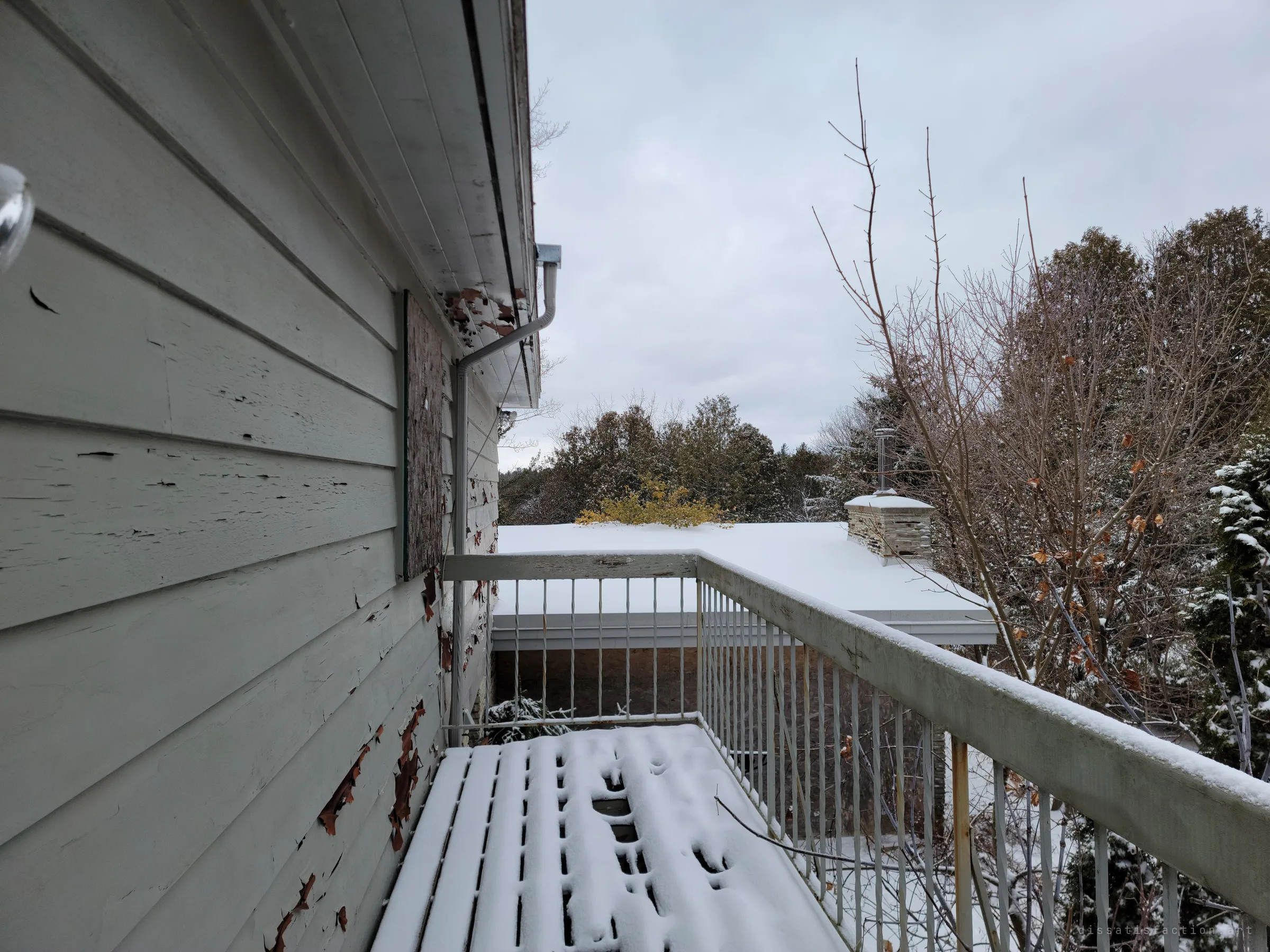 From the snowy upper balcony, peeling siding runs down to a railing that looks over a flat roof with a stone chimney and a line of evergreens. The house sits hidden in trees.