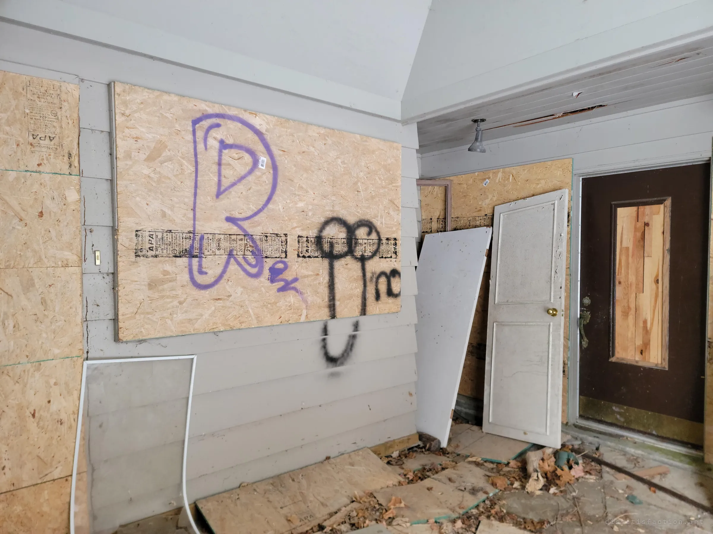Inside an enclosed porch, spray-painted tags cover a plywood-boarded window while loose doors lean against the wall. Vandals left their marks on the first room in.