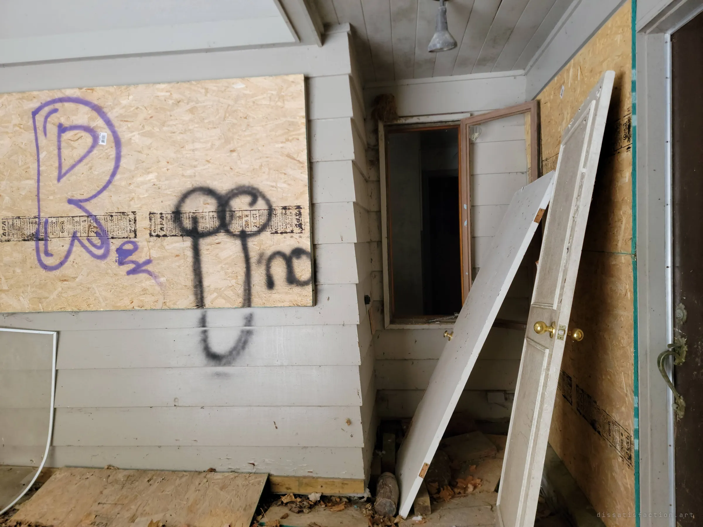 A closer look at the tagged porch wall, with an interior window hanging open into the dark house beside two doors propped against the boards.