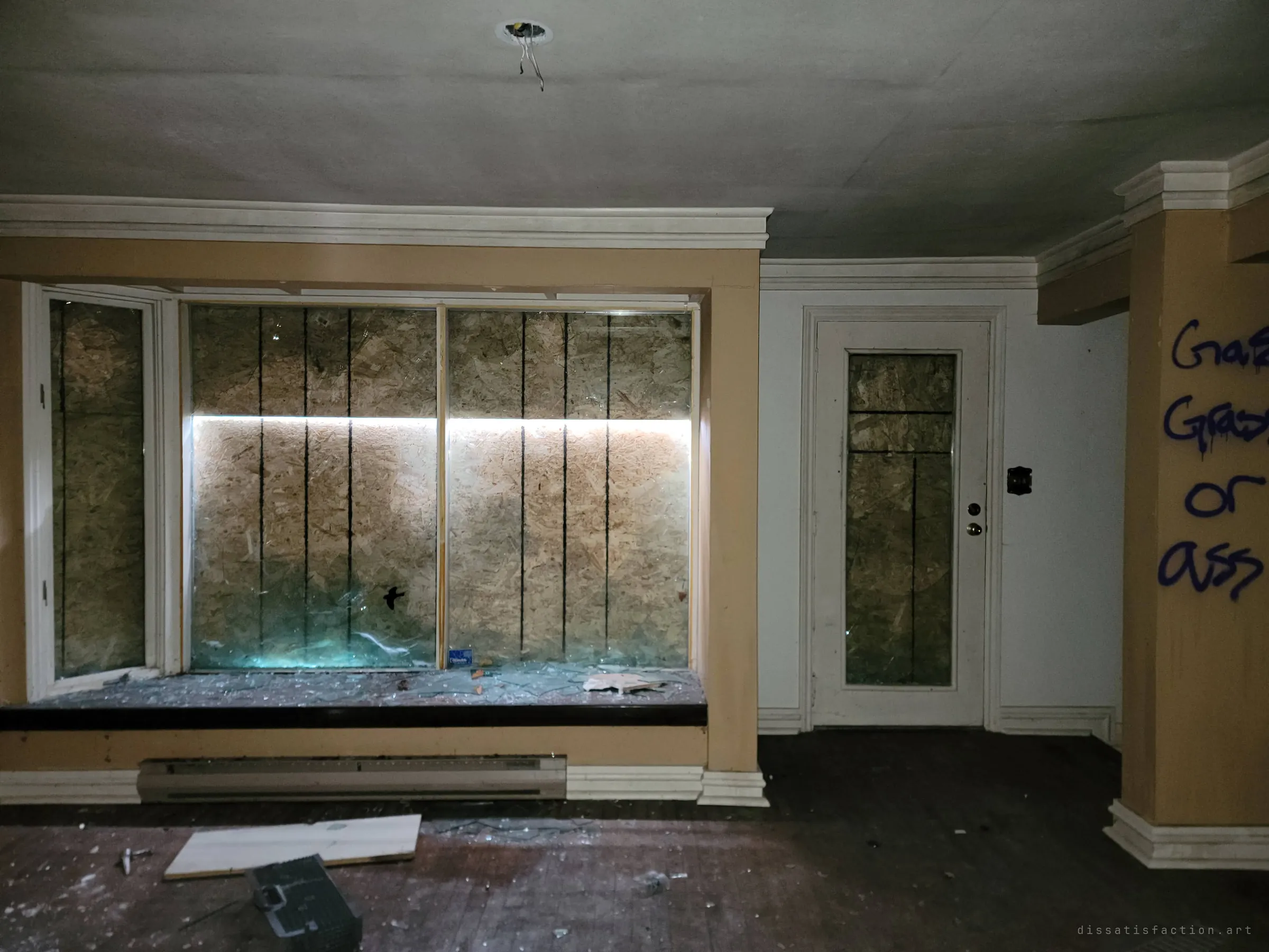 A living-room bay window, its glass shattered over the sill and the plywood behind it lit by a thin band of daylight. Crude graffiti runs down the pillar at the right.
