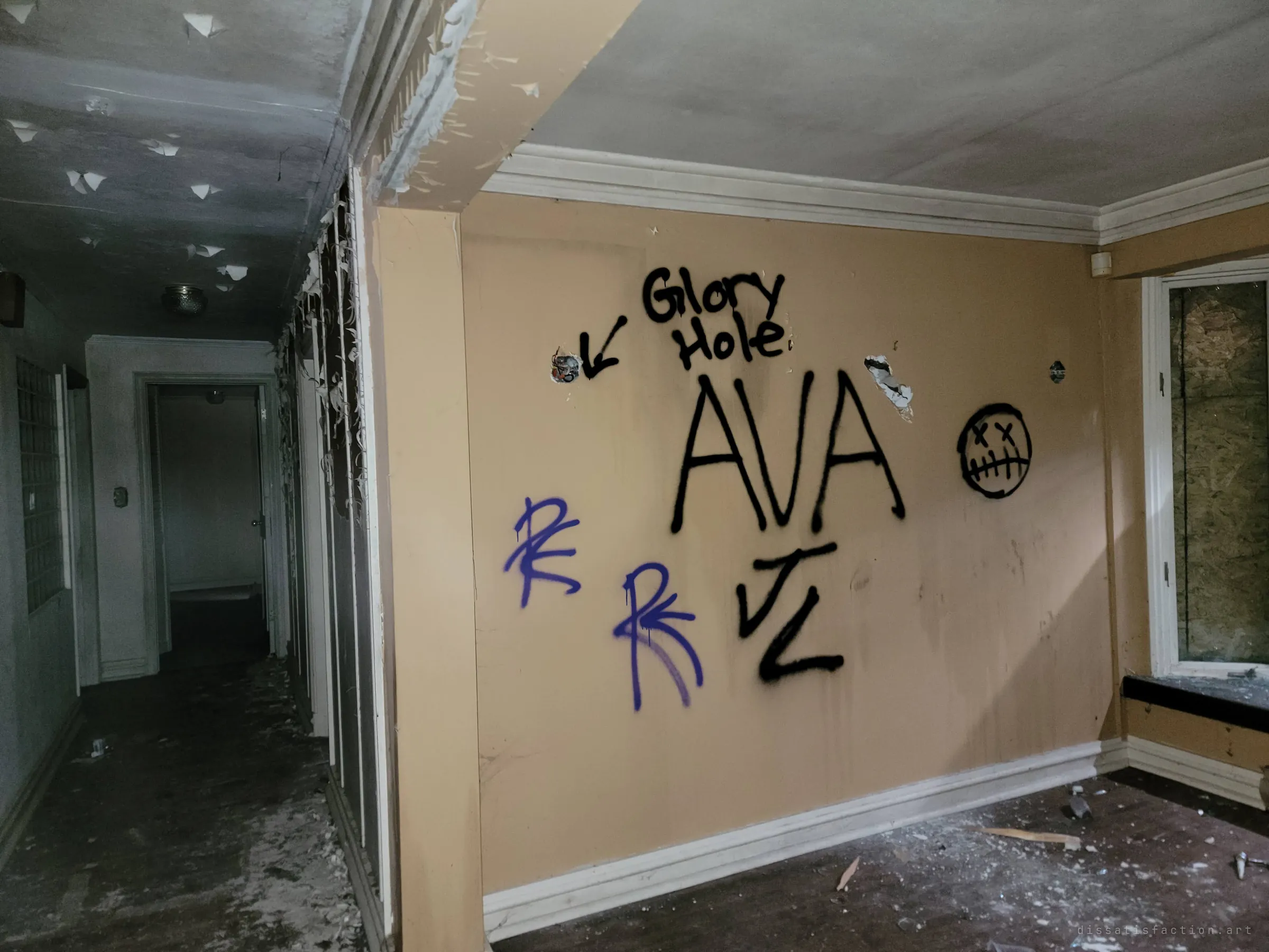 A dim hallway with the ceiling paint peeling in curls, beside a tan wall covered in spray-painted tags, a crossed-out face and a punched hole.