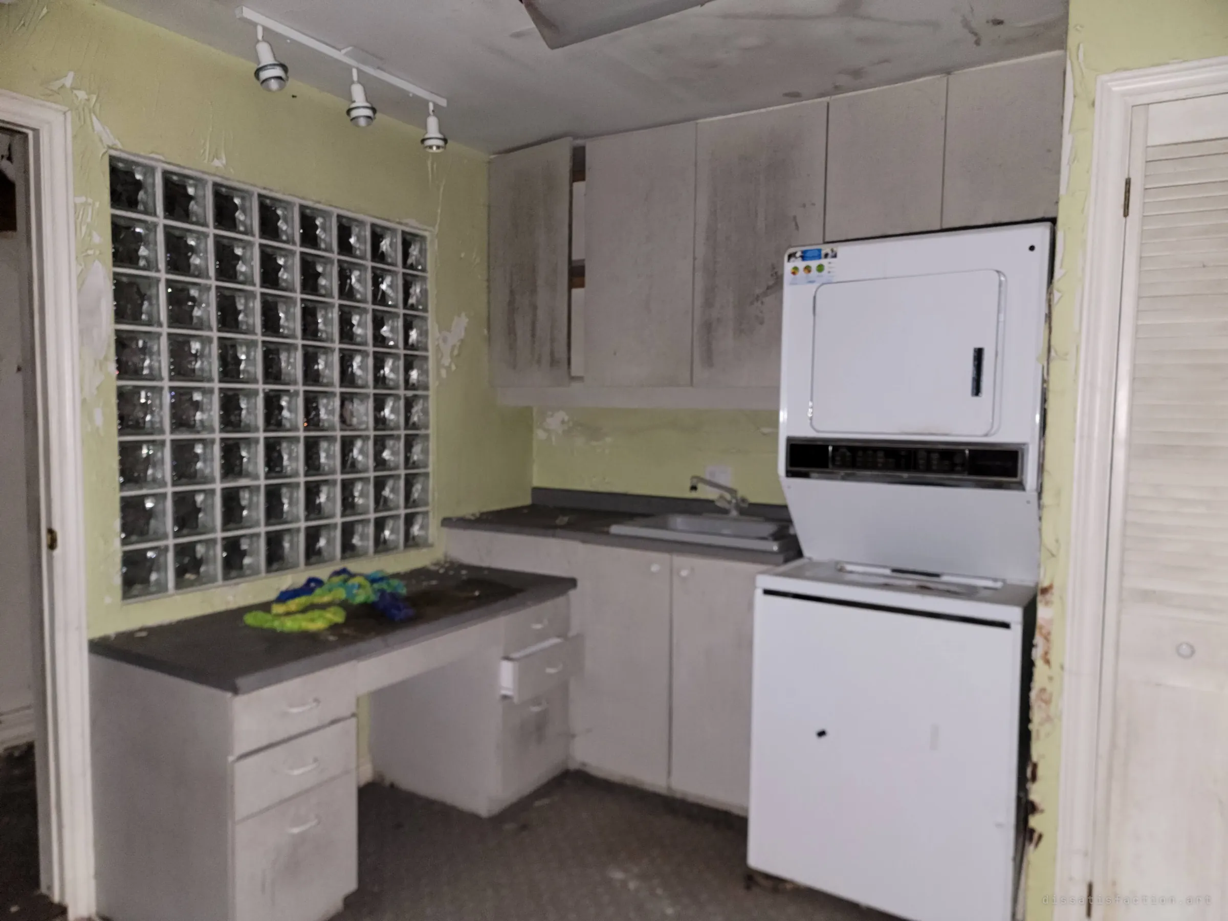 A laundry nook with a glass-block window, grimy cabinets and a stacked washer-dryer, tangled coloured rope left on the desk. The mould-streaked cabinets show how long the house has sat damp and shut.