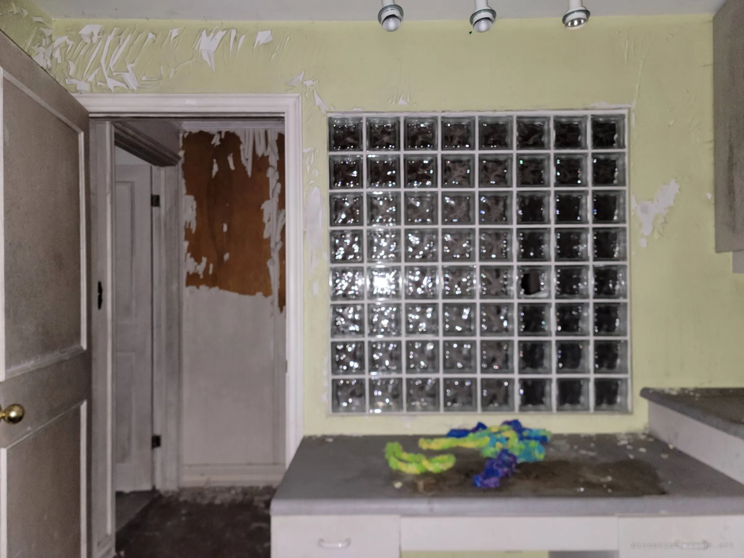 Front-on view of the glass-block wall above a dusty counter, with peeling yellow paint and a stripped doorway beside it. One broken block hints at the vandalism elsewhere in the house.