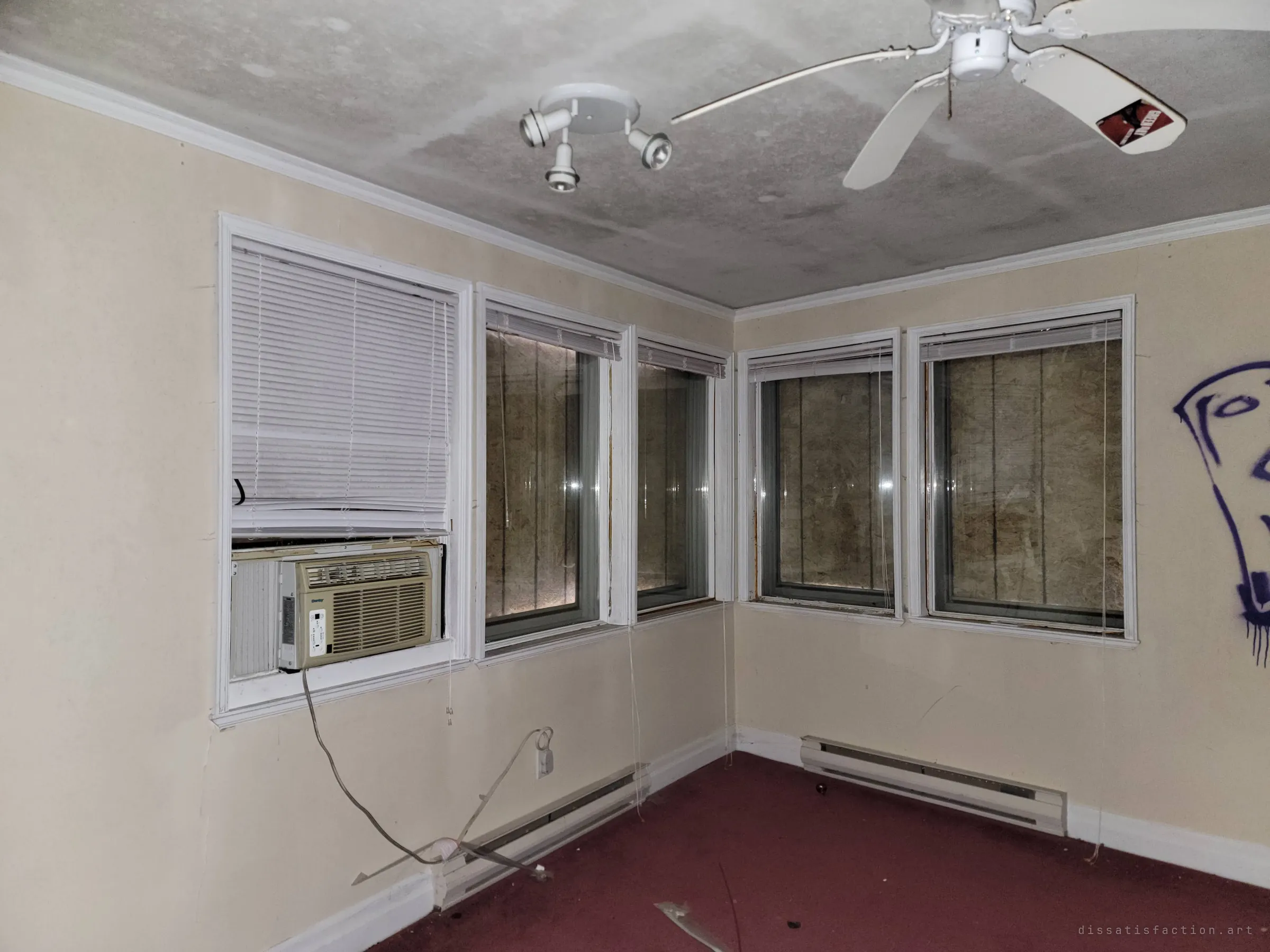 A corner room lined with boarded-over windows behind the blinds, a window air conditioner still in place and a red carpet underfoot. The plywood behind the glass shows the house was sealed up after being broken into.