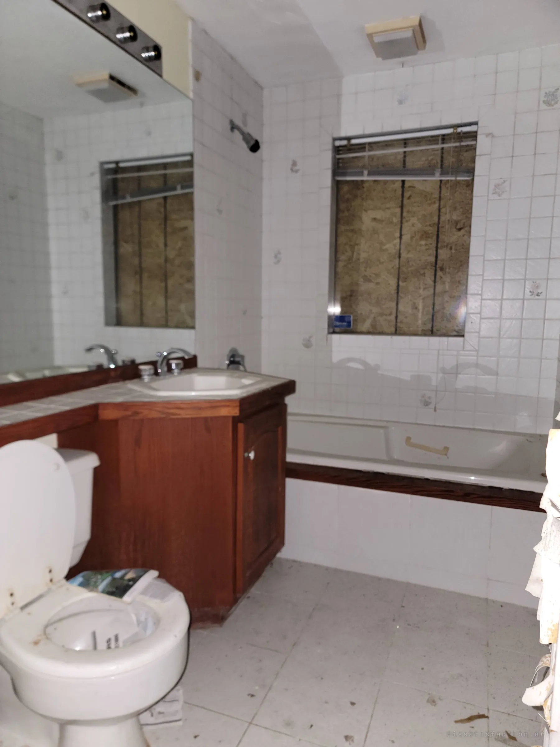 A tiled bathroom with a wooden vanity, bathtub and a boarded window, loose papers dumped on the toilet. The wall-length mirror doubles the plywood window back into the room.