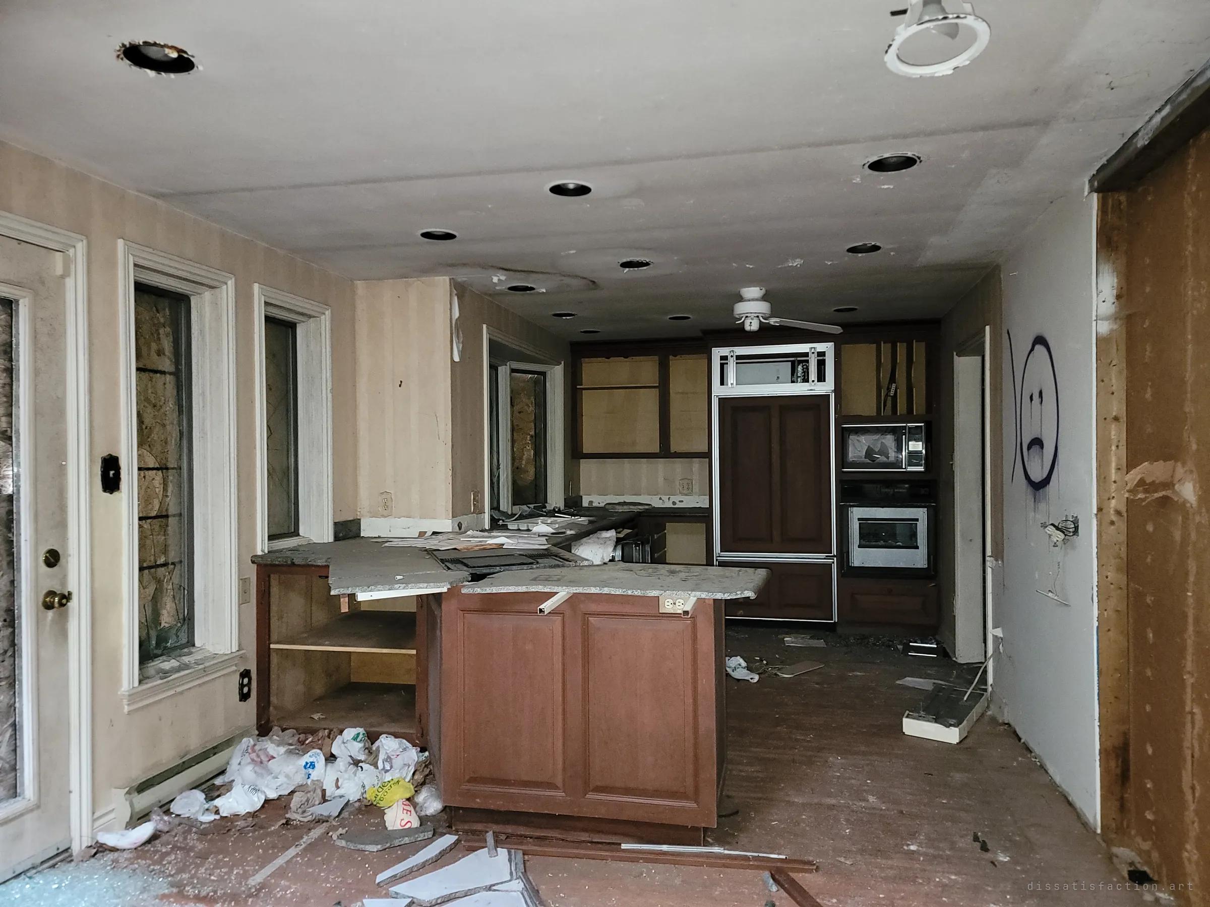 An open kitchen with a smashed stone countertop, shopping bags spilling across the floor and a sad face spray-painted on the wall. Every window along the left is boarded, leaving the room lit only by the flash.