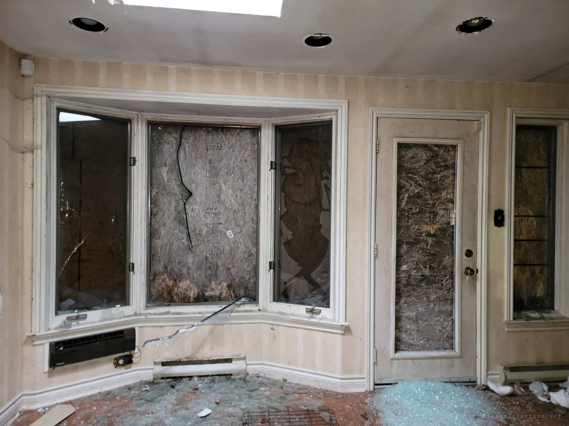 A bay window and a glass door, all their panes shattered and backed with plywood, broken glass carpeting the floor. It is the clearest record of how the vandals got in.